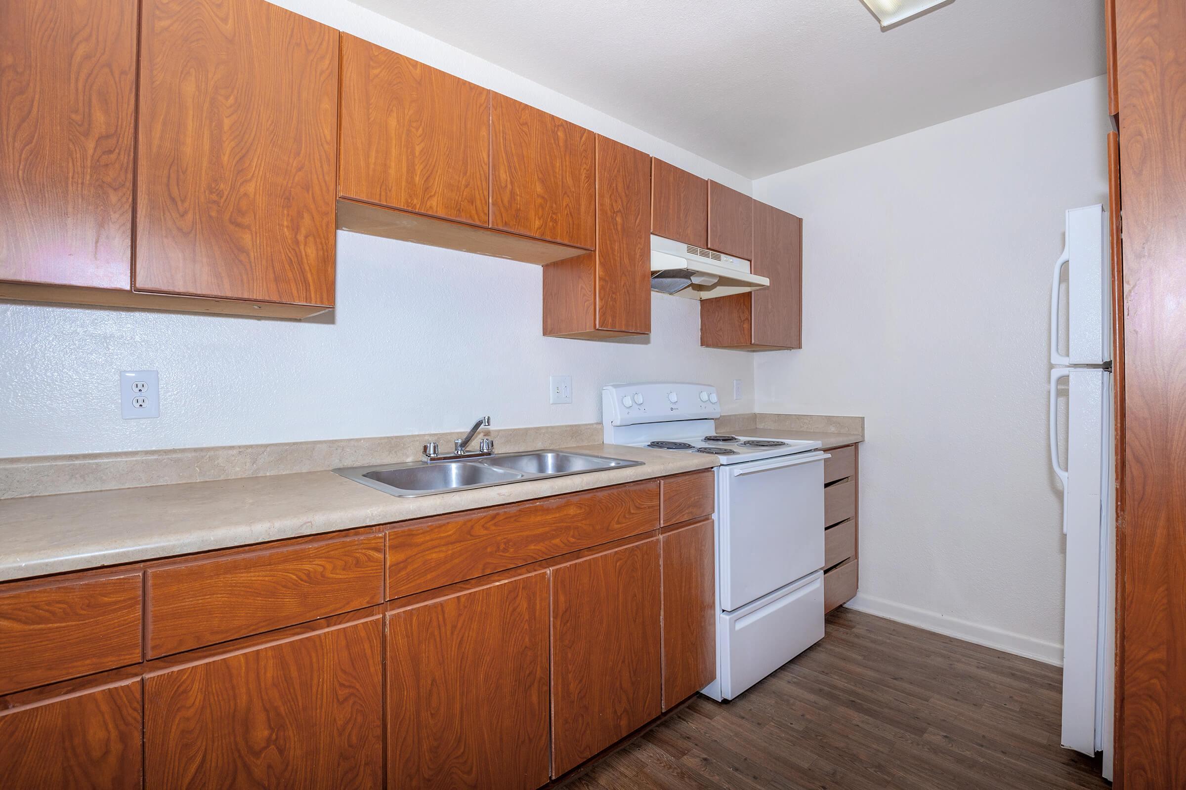 A modern kitchen featuring wooden cabinets, a countertop, a stainless steel sink, a white stove, and an oven. There is a refrigerator visible, and the flooring is a dark wood laminate. The walls are painted white, creating a bright and spacious atmosphere.