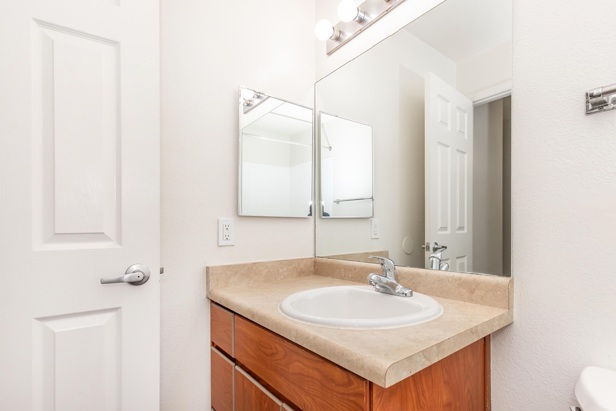 A clean bathroom with a single sink and wooden cabinetry. A large mirror is above the sink, reflecting light from wall-mounted fixtures. The walls are painted in neutral tones, and a white door leads to another room, enhancing the space's bright and spacious appearance.