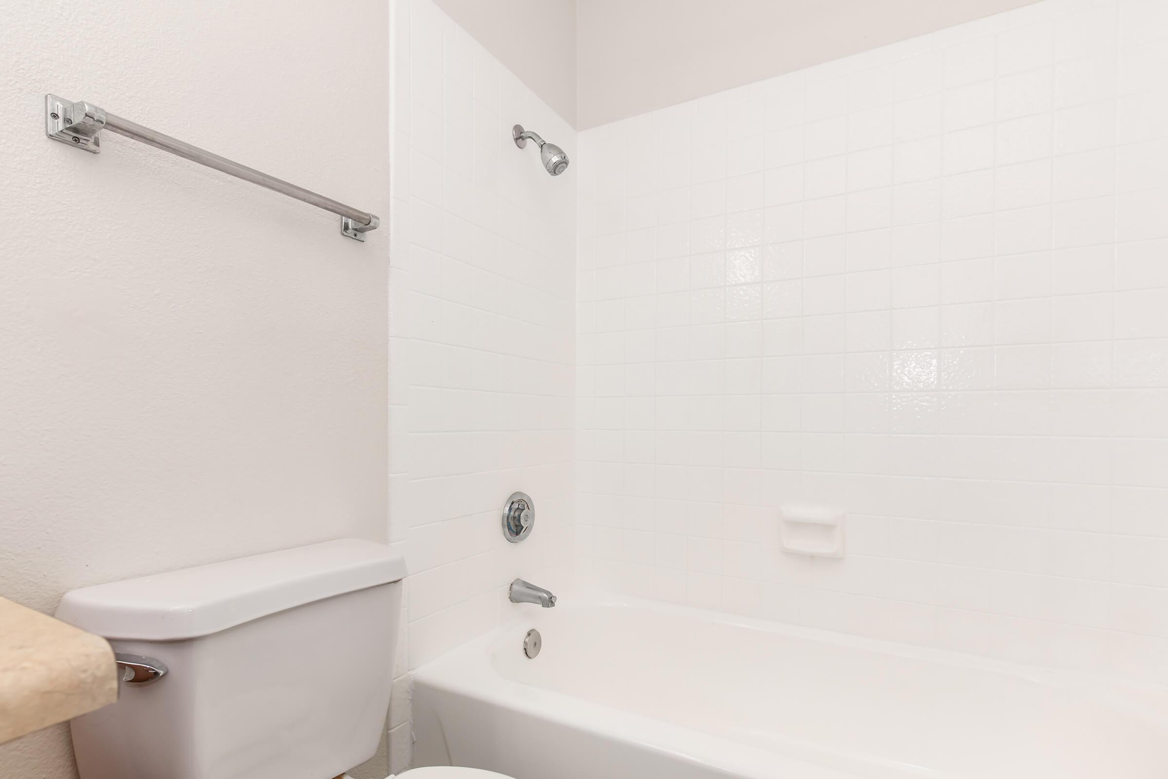 A clean bathroom featuring a white tiled wall, a bathtub, a showerhead, and a toilet. The space is well-lit with a simple design, emphasizing a minimalist and tidy look.