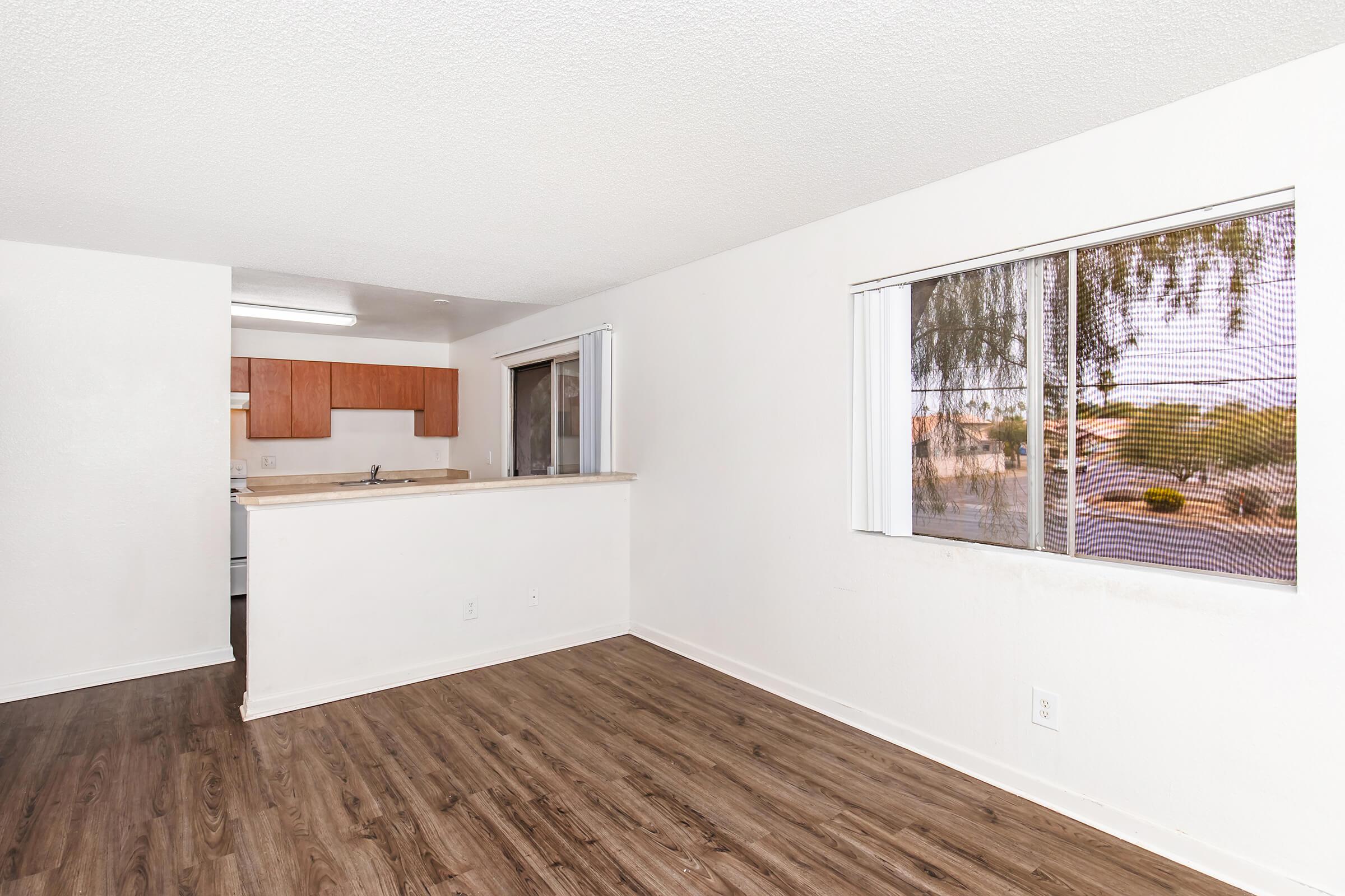 A bright, unfurnished living space featuring modern wood-like flooring, large windows allowing natural light, and a kitchen area in the background with wooden cabinets. The walls are painted white, creating a clean and open feel.