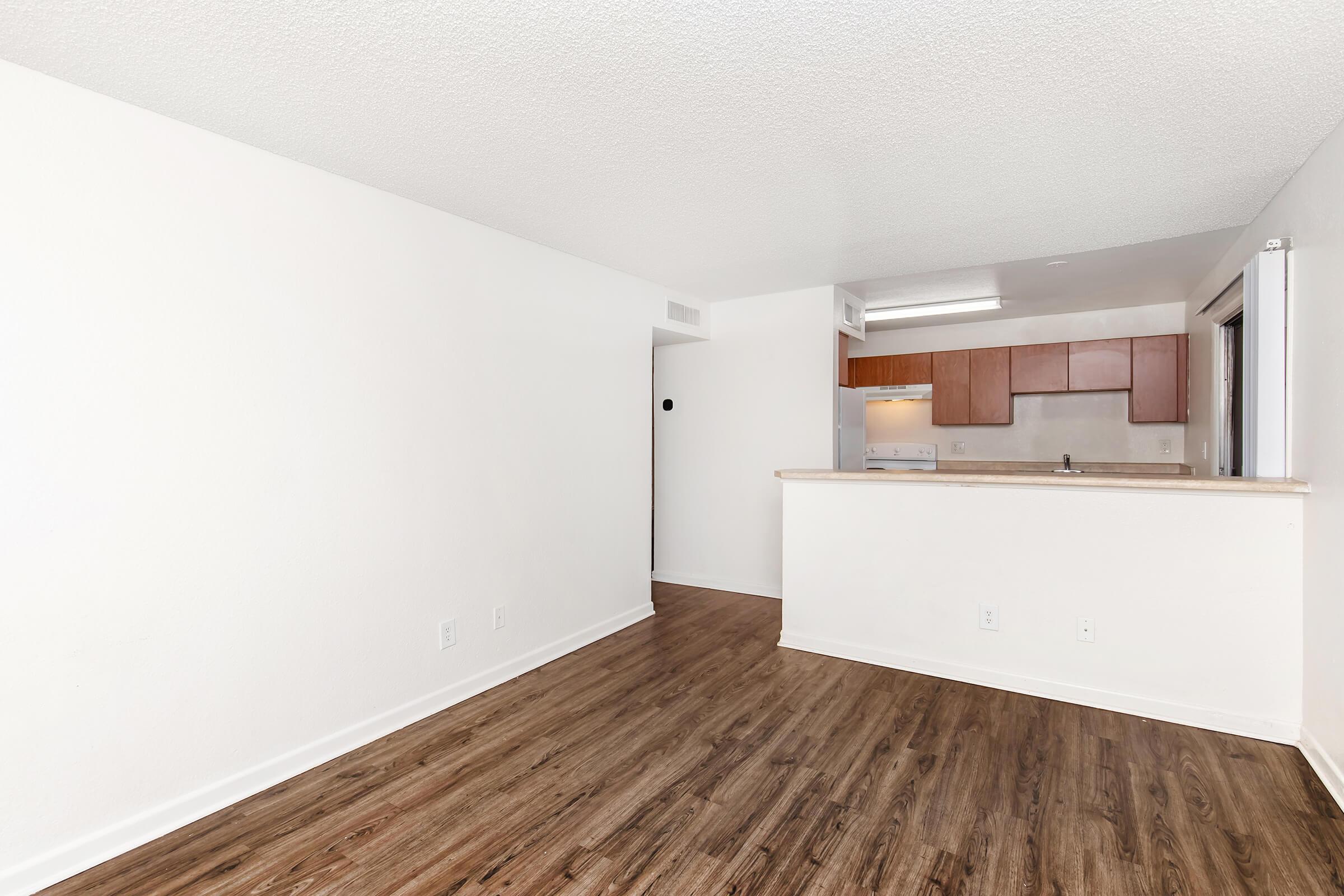 A spacious, well-lit apartment interior featuring a living area with light-colored walls and laminate flooring. In the background, there is a kitchen with wooden cabinets and a bar counter. The layout appears open and inviting, with a doorway leading into another room.