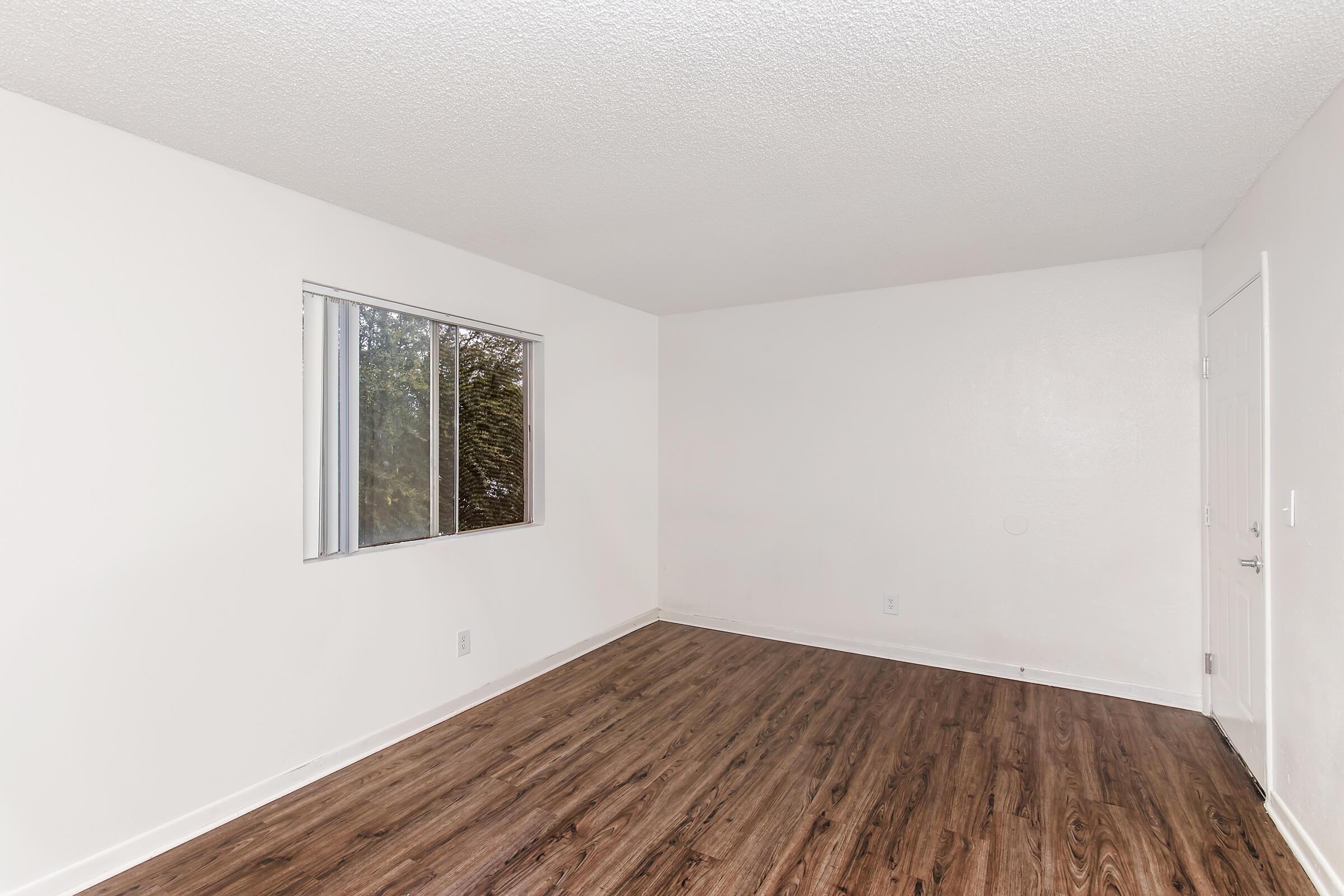 Empty room with light-colored walls and a hardwood floor. A single window provides natural light, and the room appears spacious and unfurnished. The door is visible on the right side, and there are no furniture or decorations present.