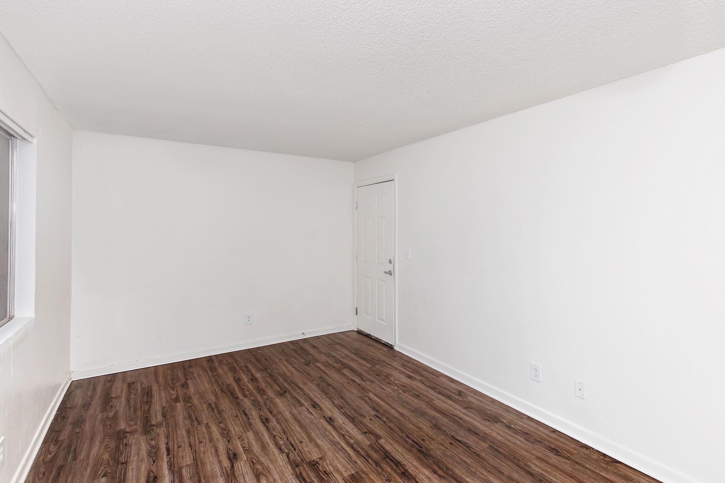 Empty room with white walls and a wooden floor. A single door is visible on the right side, and a window is on the left. The space is well-lit with natural light coming through the window, creating a simple and clean atmosphere.