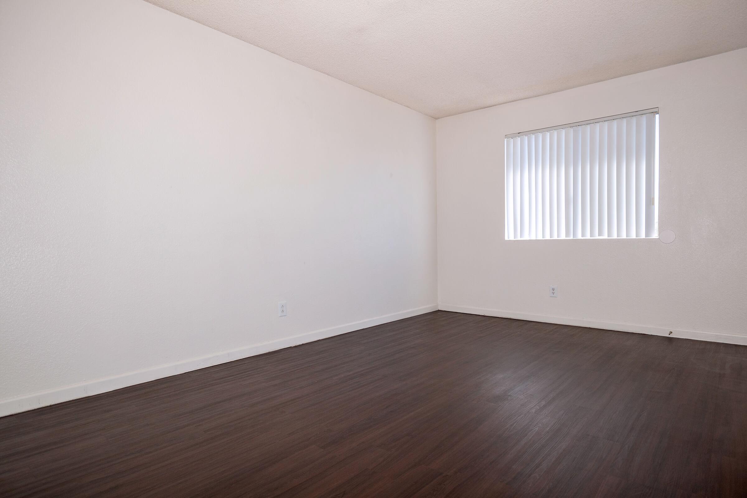 A spacious, empty room with light brown laminate flooring and white walls. A small window with vertical blinds is visible on one wall, allowing natural light to enter. The room has no furniture, creating a clean and minimalistic appearance.