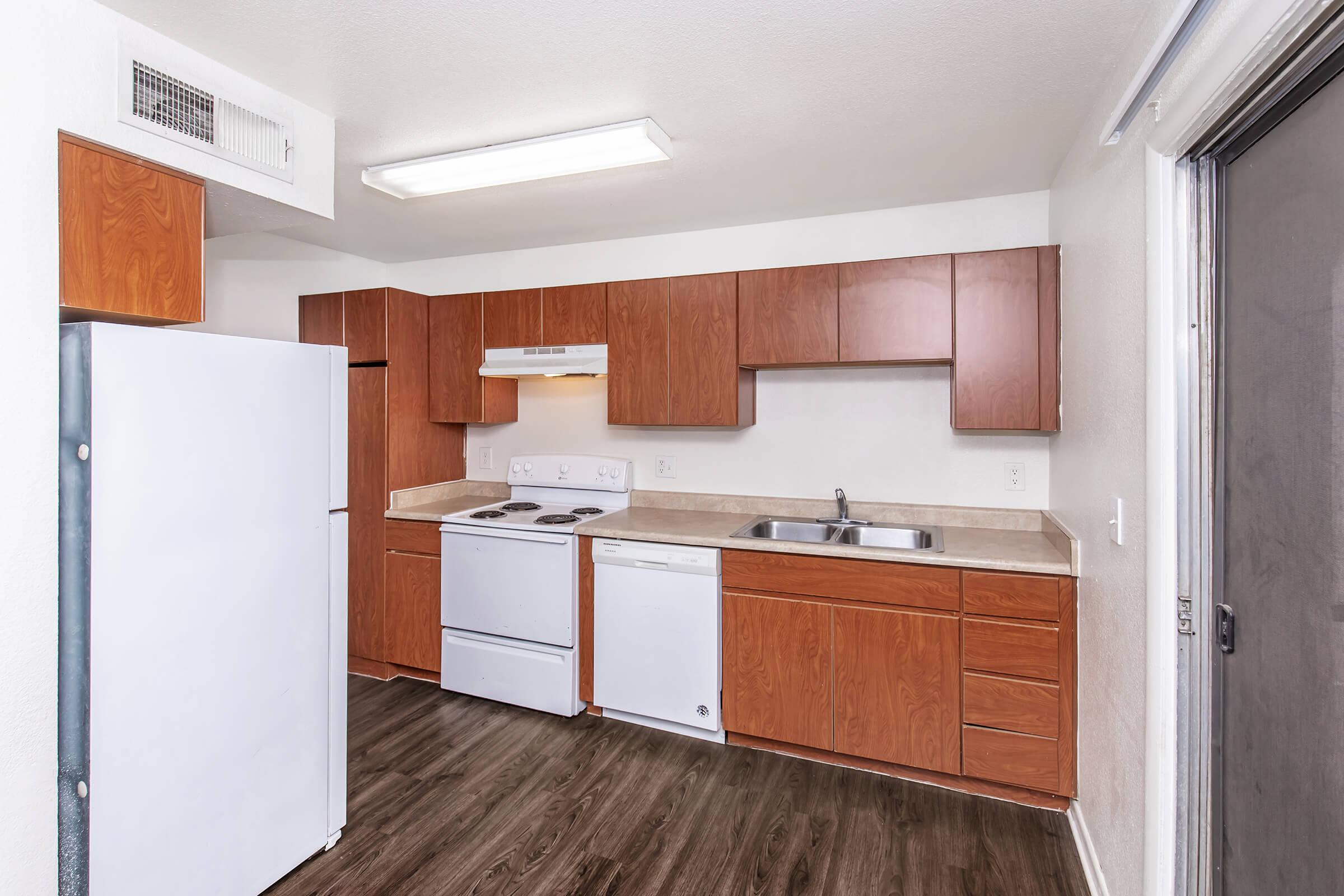 A modern kitchen featuring wooden cabinetry, a white refrigerator, a white stove, and a dishwasher. The countertop is beige, and the flooring is dark wood. There is a single basin sink under a wall-mounted range hood, with a ceiling light illuminating the space.