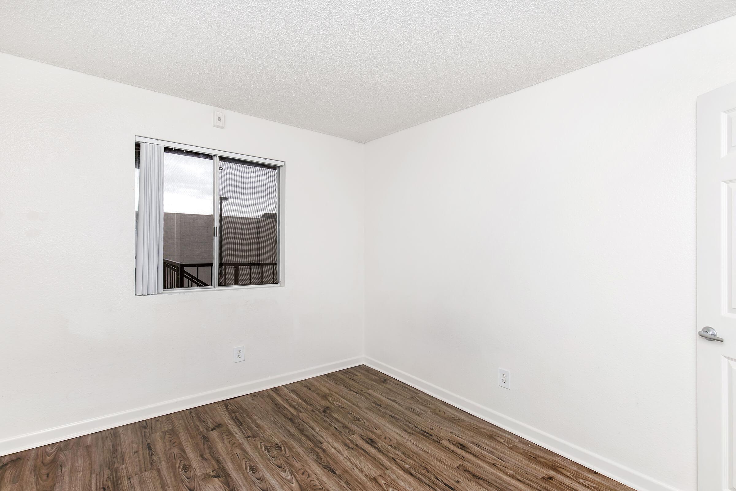 Empty room with light-colored walls and a wood-like laminate floor. A window with a sheer curtain allows natural light to enter. The space is minimally furnished, creating a clean and open atmosphere.