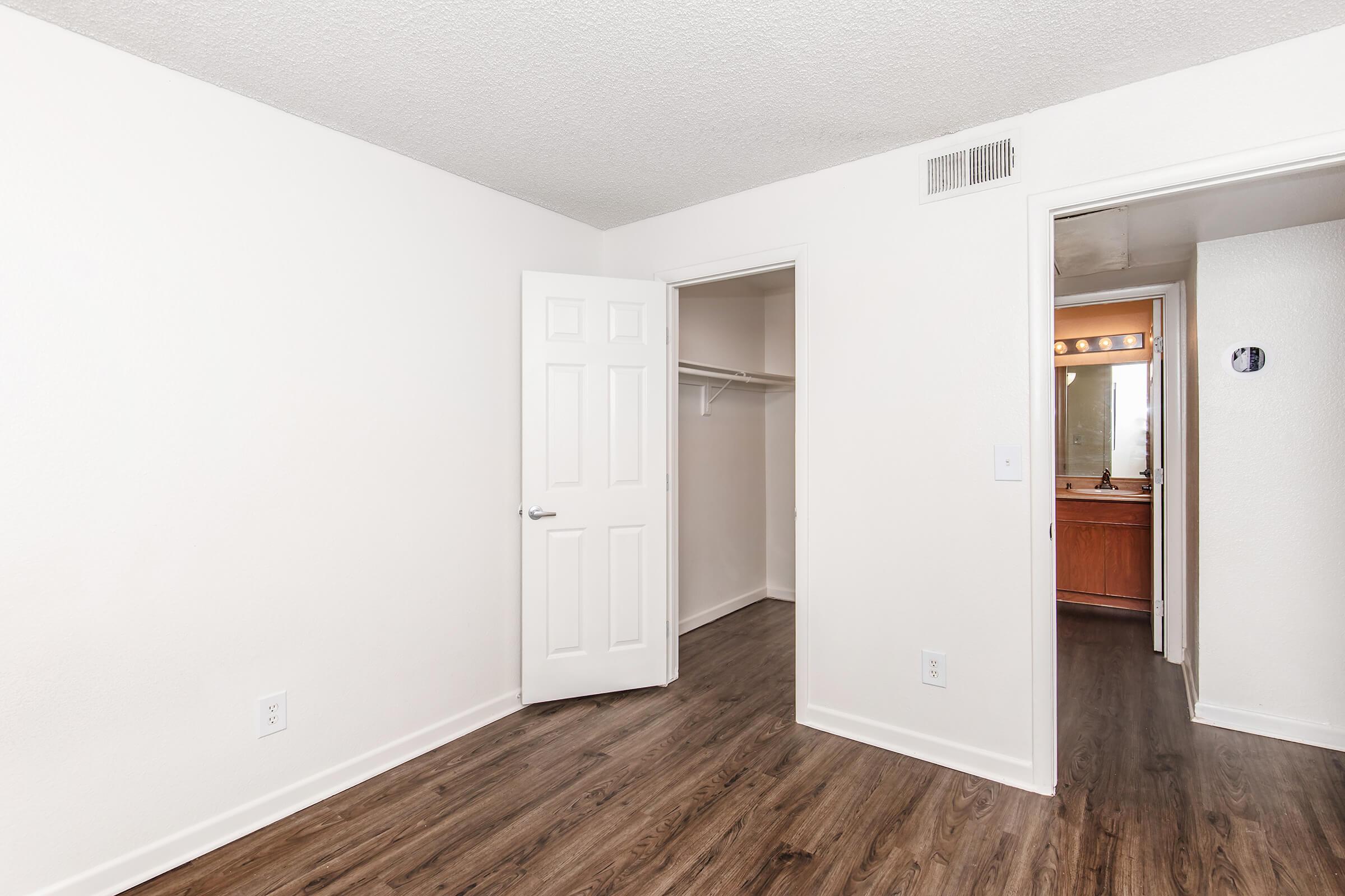 A well-lit, empty room featuring light-colored walls and a smooth wooden floor. There is a white closet door on the left and an open door leading to a bathroom on the right. The room is simple and spacious, ideal for personal decoration or furnishing.