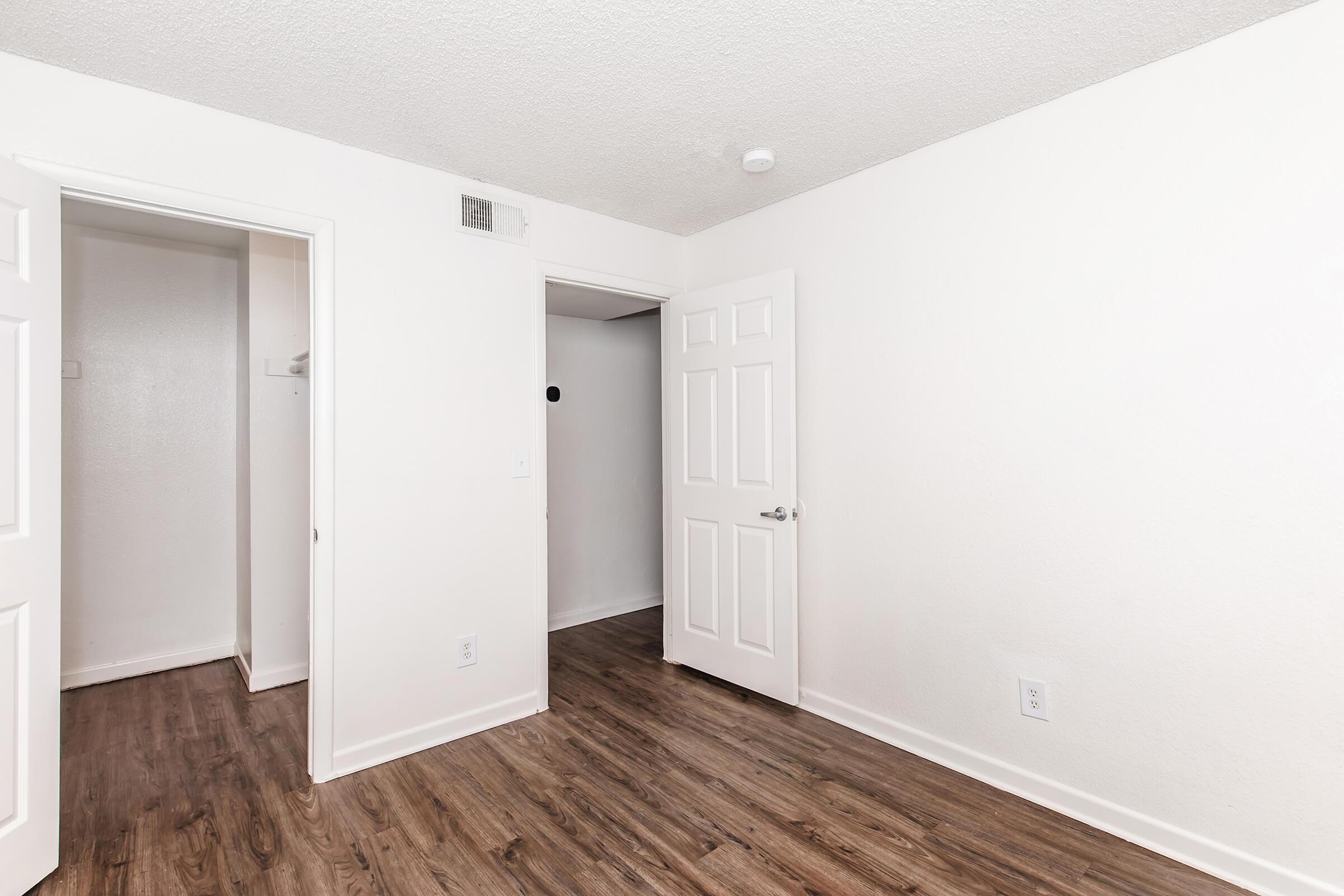 An empty room with light-colored walls and a wood-like floor. There are two open doors, one leading to a small closet and the other to an adjacent space. The room is well-lit and has a simple, minimalist design with no furniture or decorations.