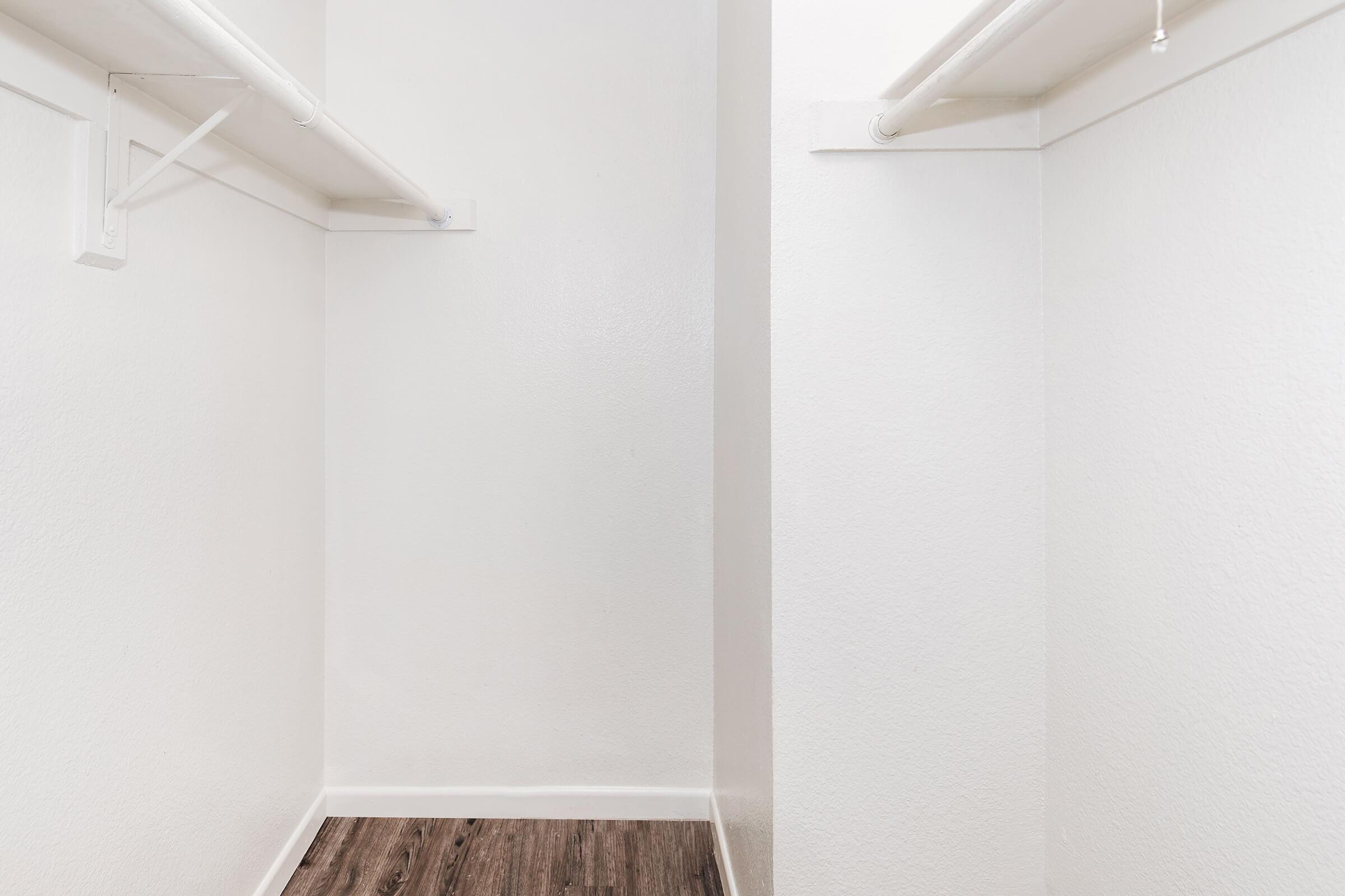Empty closet with white-painted walls and two hanging rods, one on each wall. The floor has a dark wood-like finish, creating a contrasting look. The space is well-lit and minimalist in design, emphasizing the openness and potential for storage.