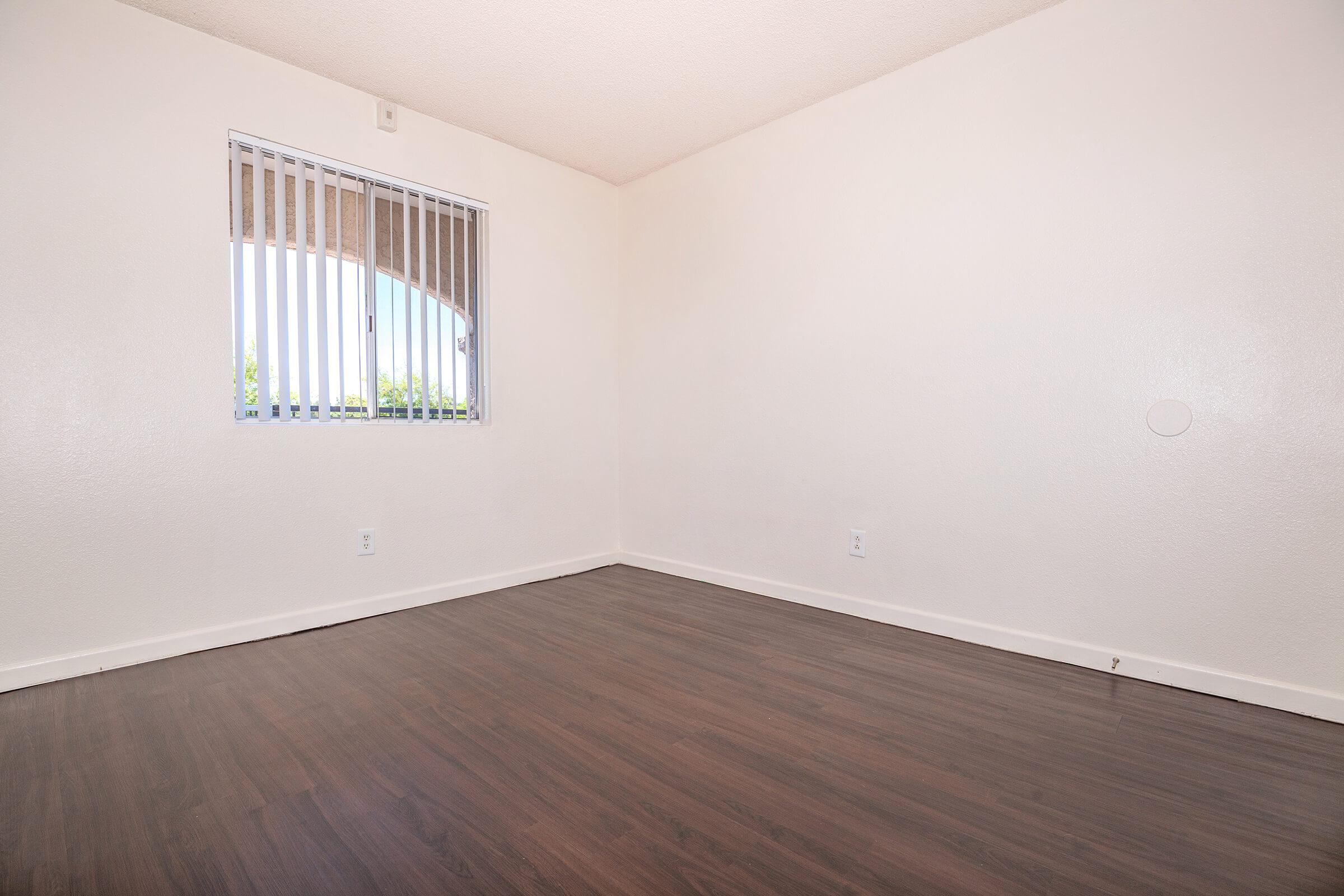A spacious, empty room featuring light-colored walls and a wood laminate floor. A window with vertical blinds allows natural light to enter, while the room remains unfurnished, emphasizing its openness and potential for personalization.