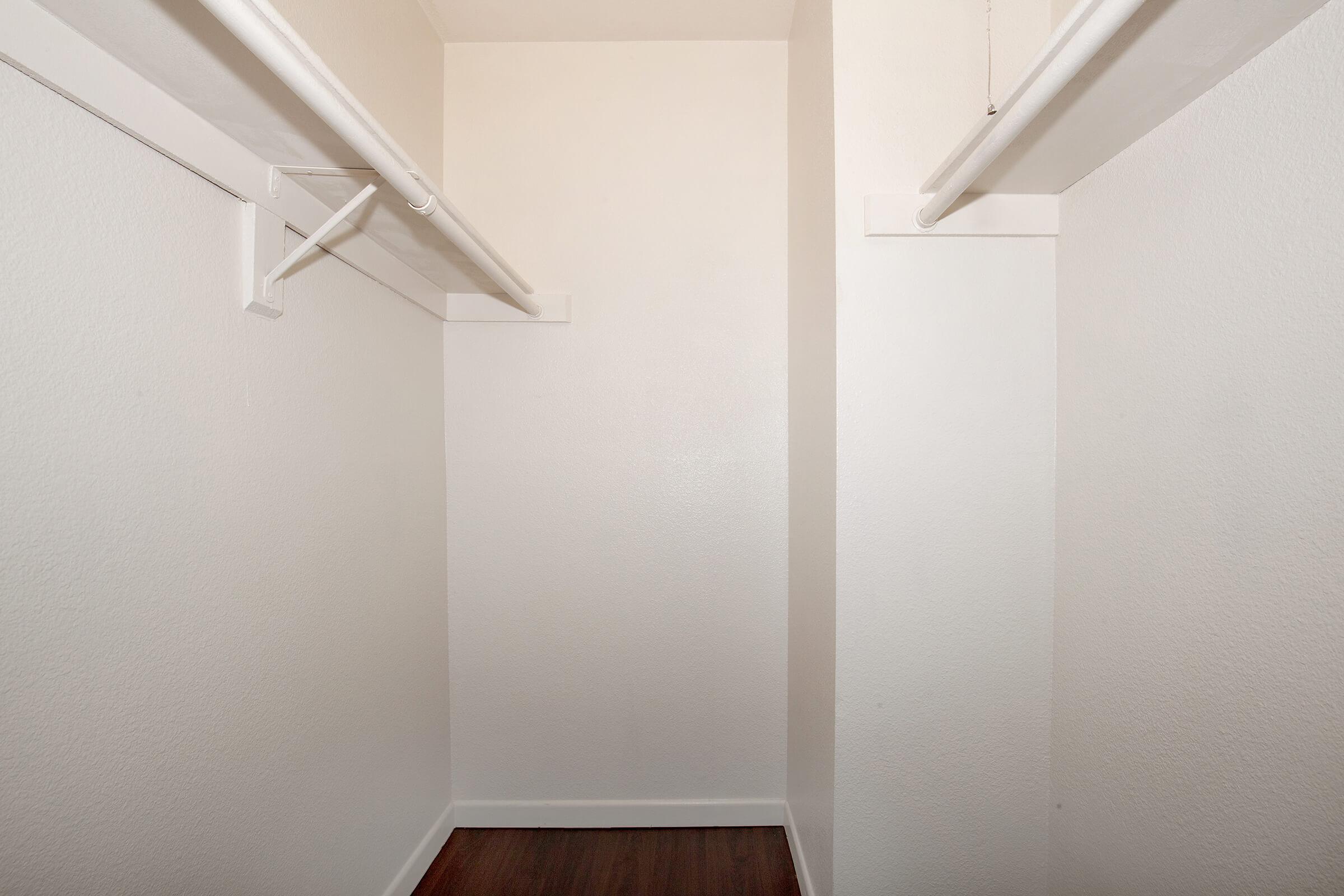 Empty walk-in closet with white walls, two hanging rods along the sides, and wooden flooring. The space is well-lit and uncluttered, creating a minimalist aesthetic.