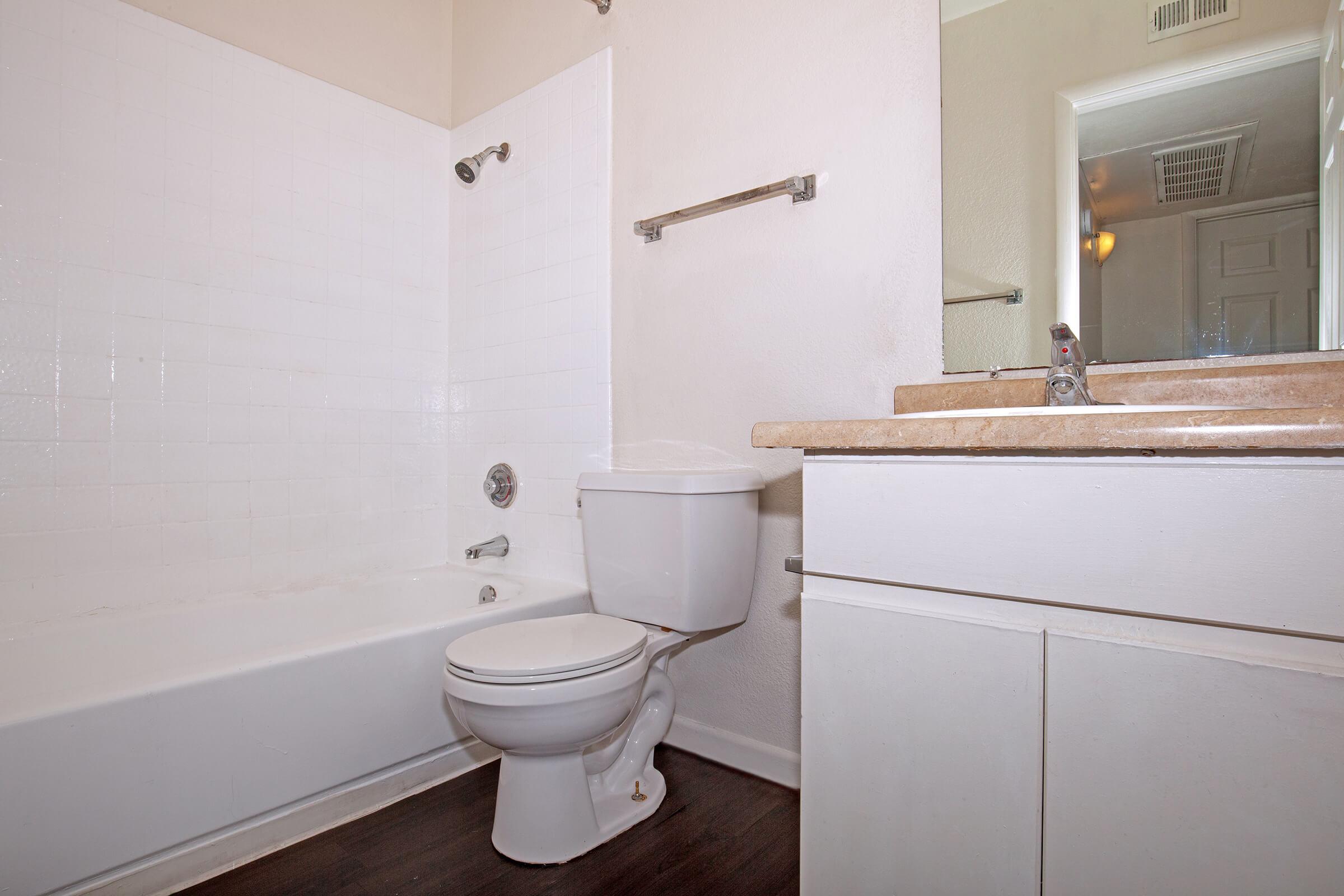 A clean, modern bathroom featuring a white bathtub with shower, a toilet, and a stylish sink with a countertop. There is a mirror above the sink, and the walls are painted in a light color, creating a bright and open atmosphere. The flooring is dark, providing a contrast to the lighter elements.