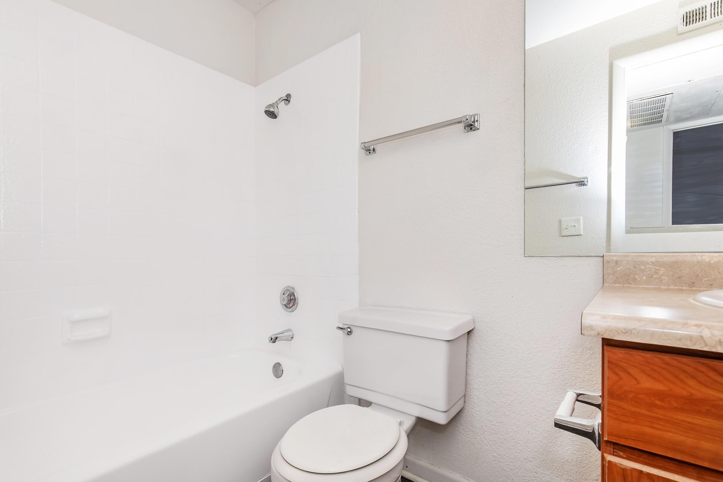 A clean and simple bathroom featuring a white bathtub surrounded by tiled walls, a toilet, a small bathroom mirror, and a countertop with wooden cabinetry. Bright lighting enhances the neutral color scheme.