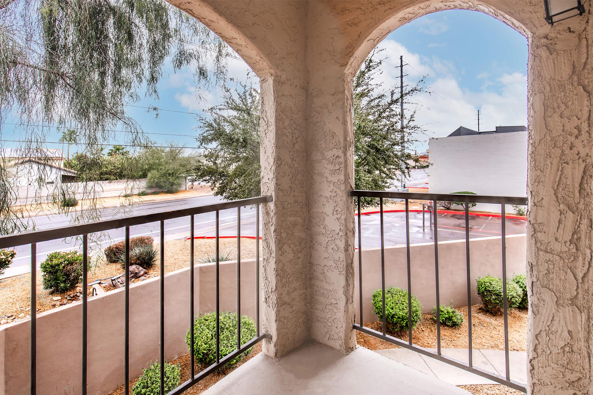 View from a balcony featuring decorative metal railings, overlooking a landscaped area with shrubs and gravel. A street and buildings are visible in the background under a partly cloudy sky. The setting suggests a suburban environment with a mix of greenery and paved surfaces.
