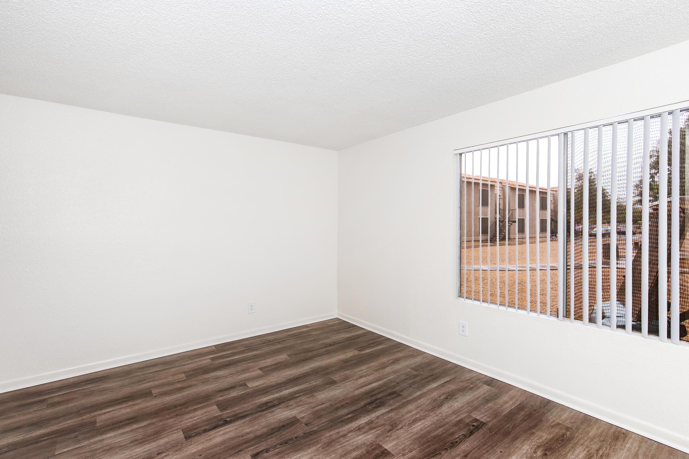 A vacant room with light-colored walls, laminate flooring, and a window featuring vertical blinds. Outside the window, there is a view of a grassy area and buildings. The room is empty, creating a spacious and airy feel.