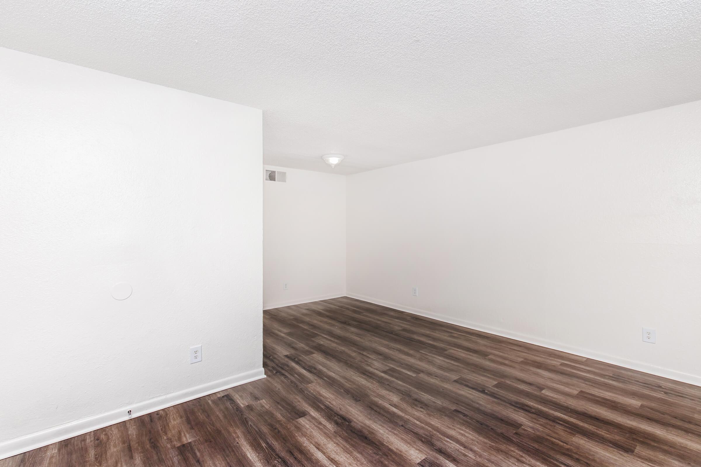 A bright, empty room featuring light-colored walls and a textured ceiling. The floor is covered in wood-like laminate, and the space is well-lit by a ceiling fixture. There are no furnishings, giving a sense of openness. The room's layout includes a corner wall separating it from another space.