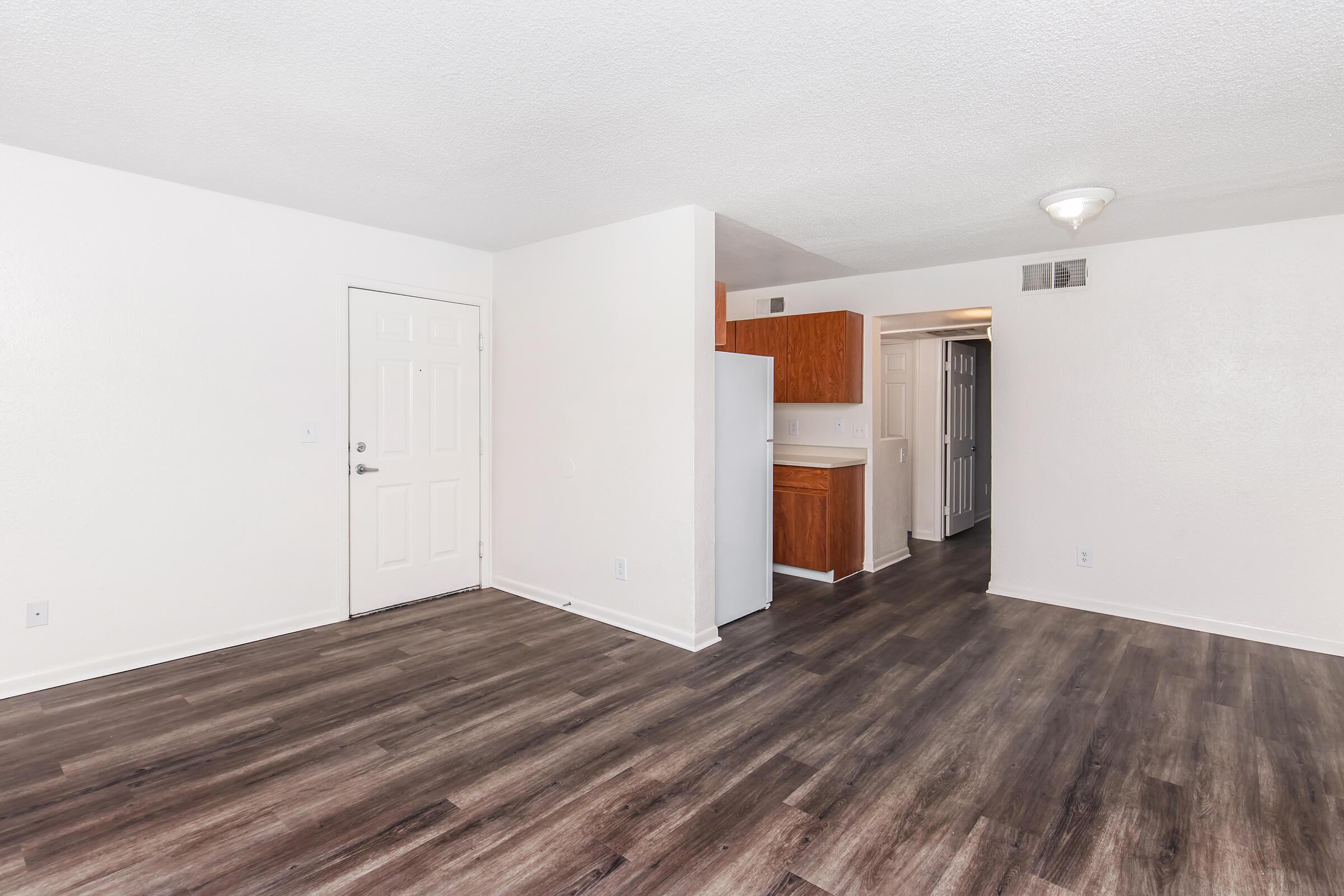 Interior view of a modern, empty living space featuring light-colored walls, a front door, and an open layout. The floor has dark wood-like laminate, and there's a small kitchenette area visible in the background. The space appears clean and well-lit, suitable for various furniture arrangements.