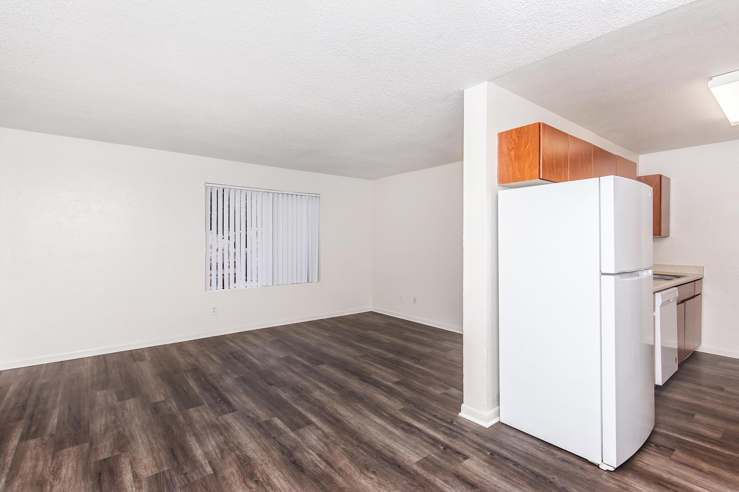 An empty, well-lit apartment interior featuring a white refrigerator and wooden cabinets. The floor has dark laminate planks, and there is a window with vertical blinds on one wall. The walls are painted a light color, creating a spacious and clean atmosphere.