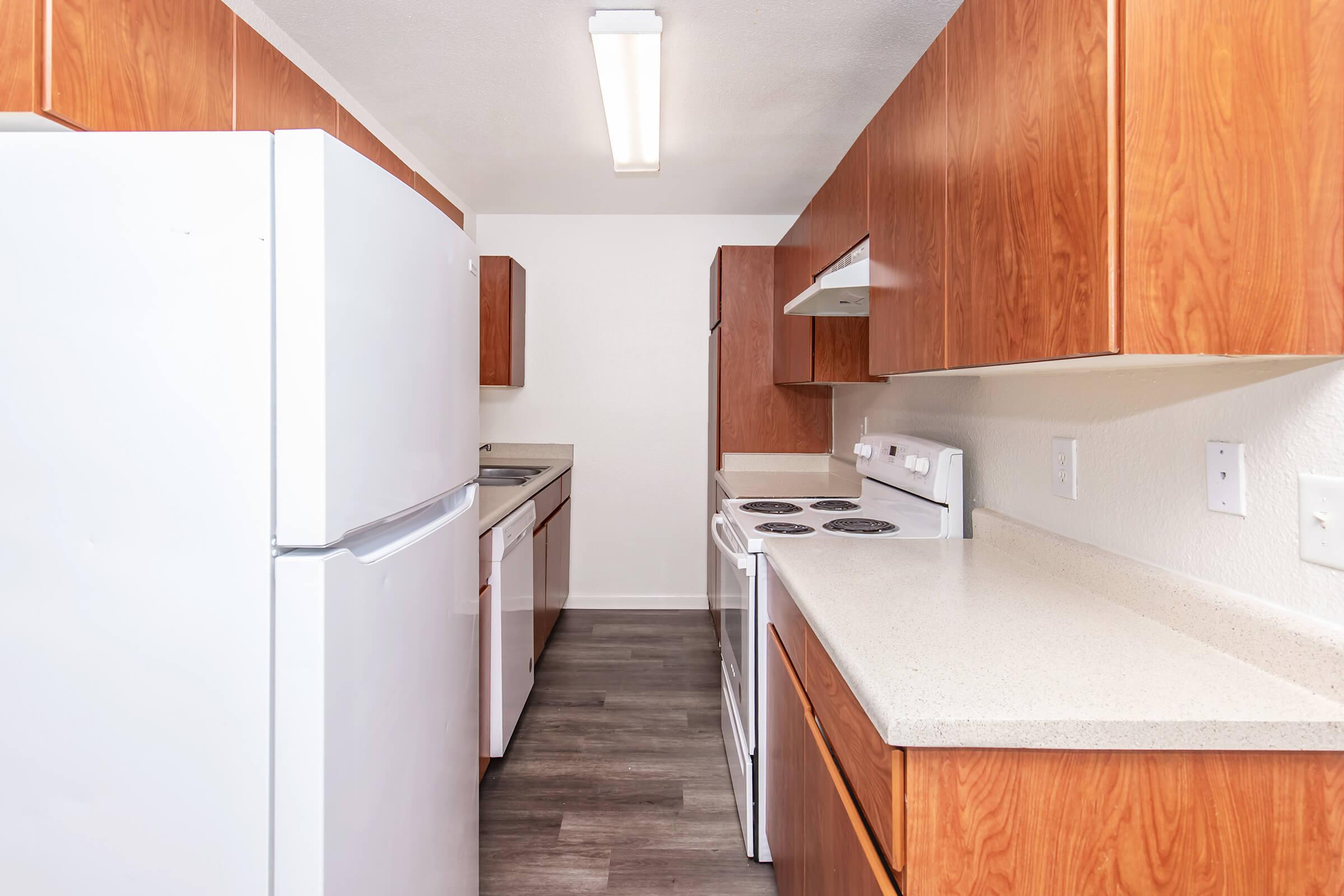 Bright kitchen with modern appliances, including a white refrigerator, stove, and microwave. The cabinetry is made of warm wood, and the countertops are light-colored. Bright overhead lighting illuminates the space, which features a clean, contemporary design with gray flooring.