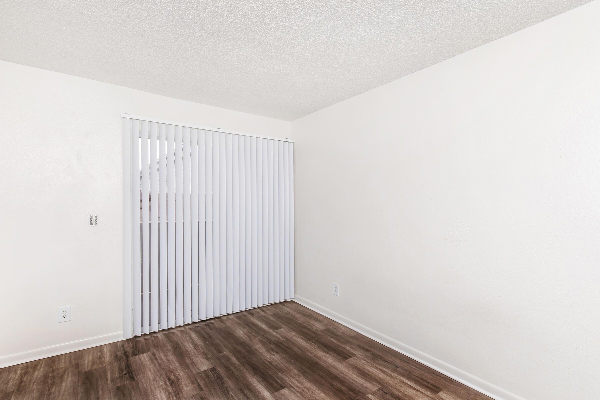 Empty room with light-colored walls and a wooden floor. A vertical blind covers a window, letting in natural light. The space is simple and clean, featuring no furniture or decorations.