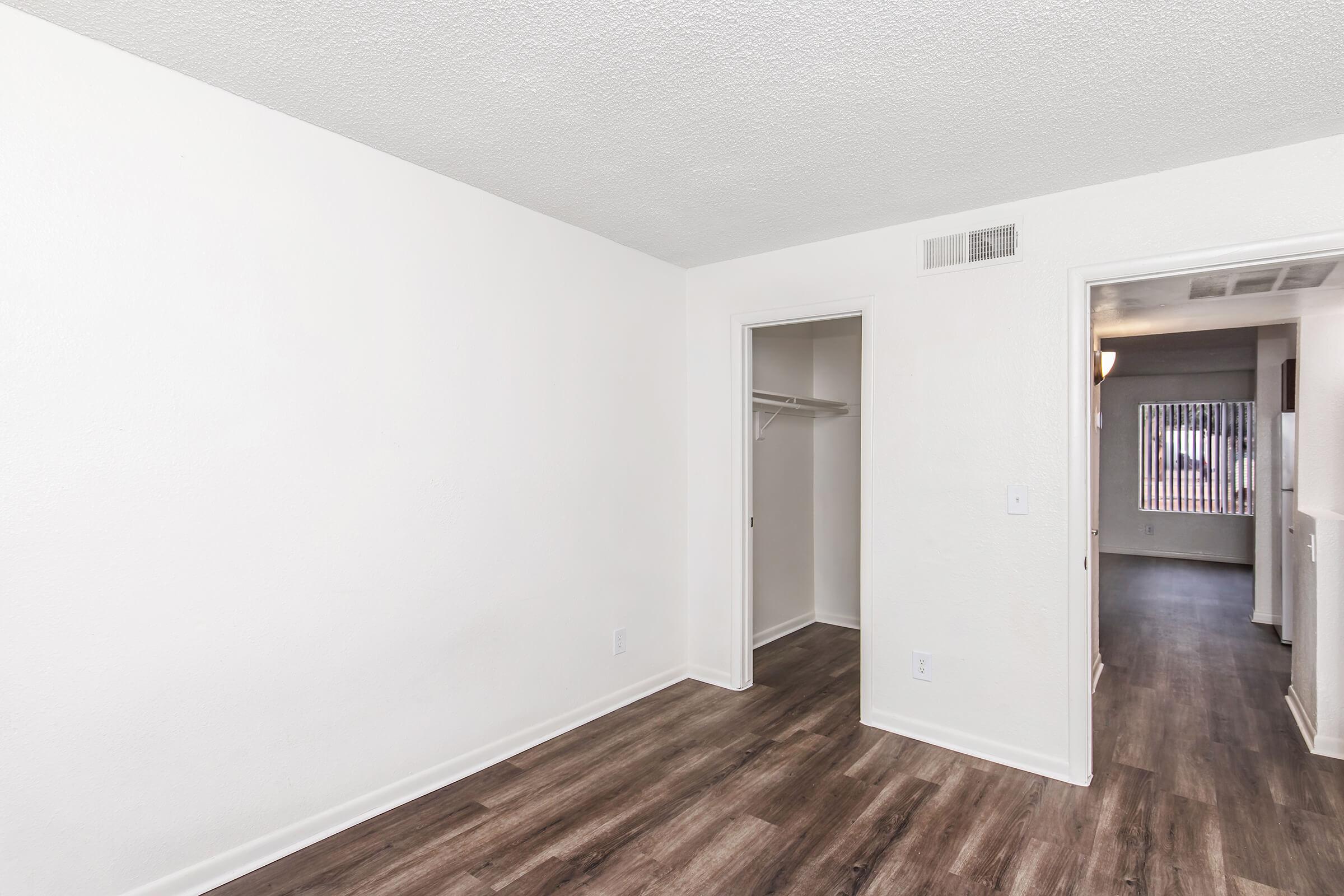 A spacious, unfurnished room with light-colored walls and a textured ceiling. The flooring is dark laminate. There is a doorway leading to another room and a small closet on the left side. Natural light enters through a window in the adjacent room, creating a bright atmosphere.