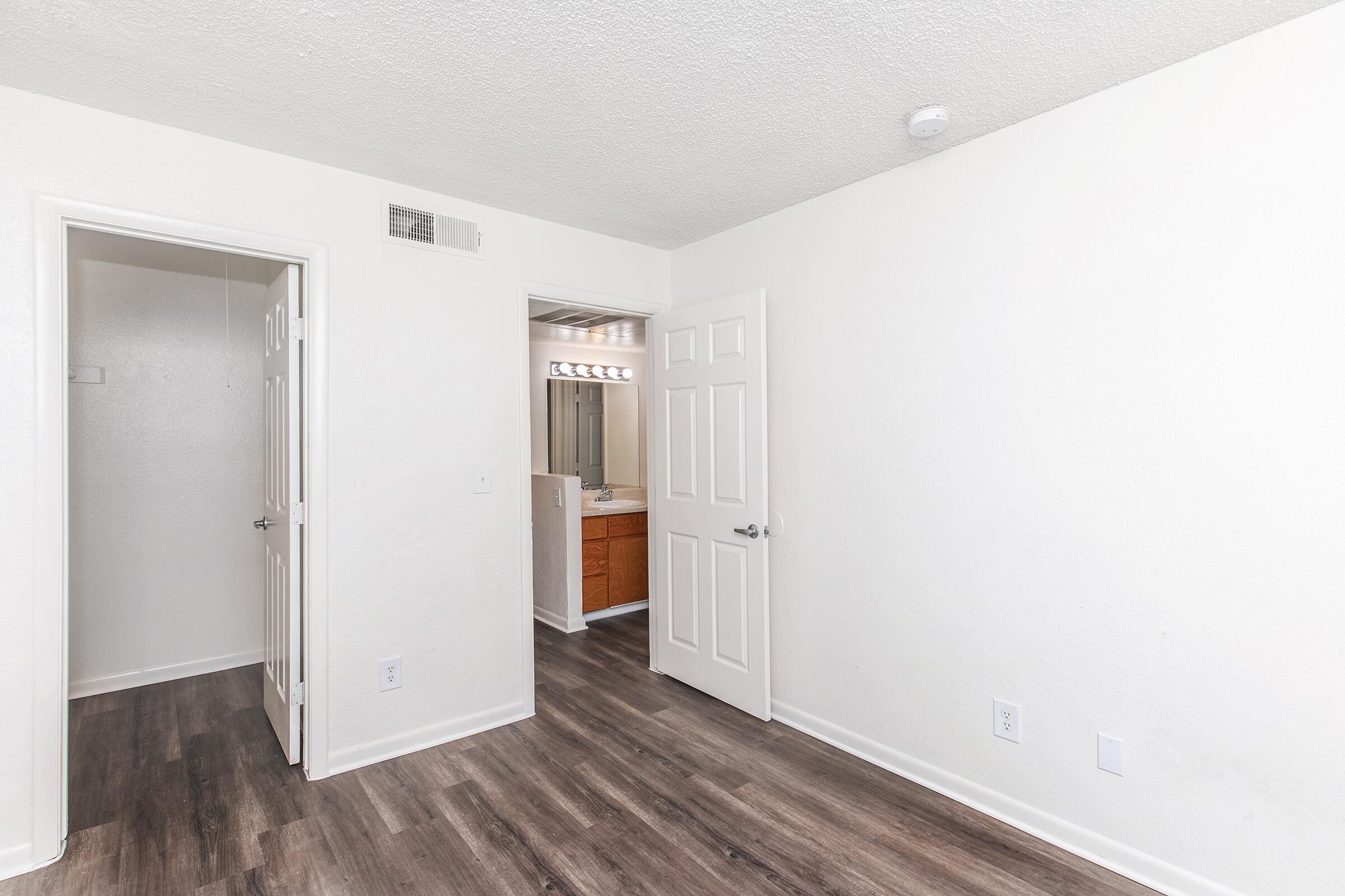 A bright, empty room with light-colored walls and wood-look flooring. A doorway leads to another room on the left, while another door on the right opens to a bathroom with vanity lighting. The room features a ceiling with a light fixture and a wall-mounted smoke detector.