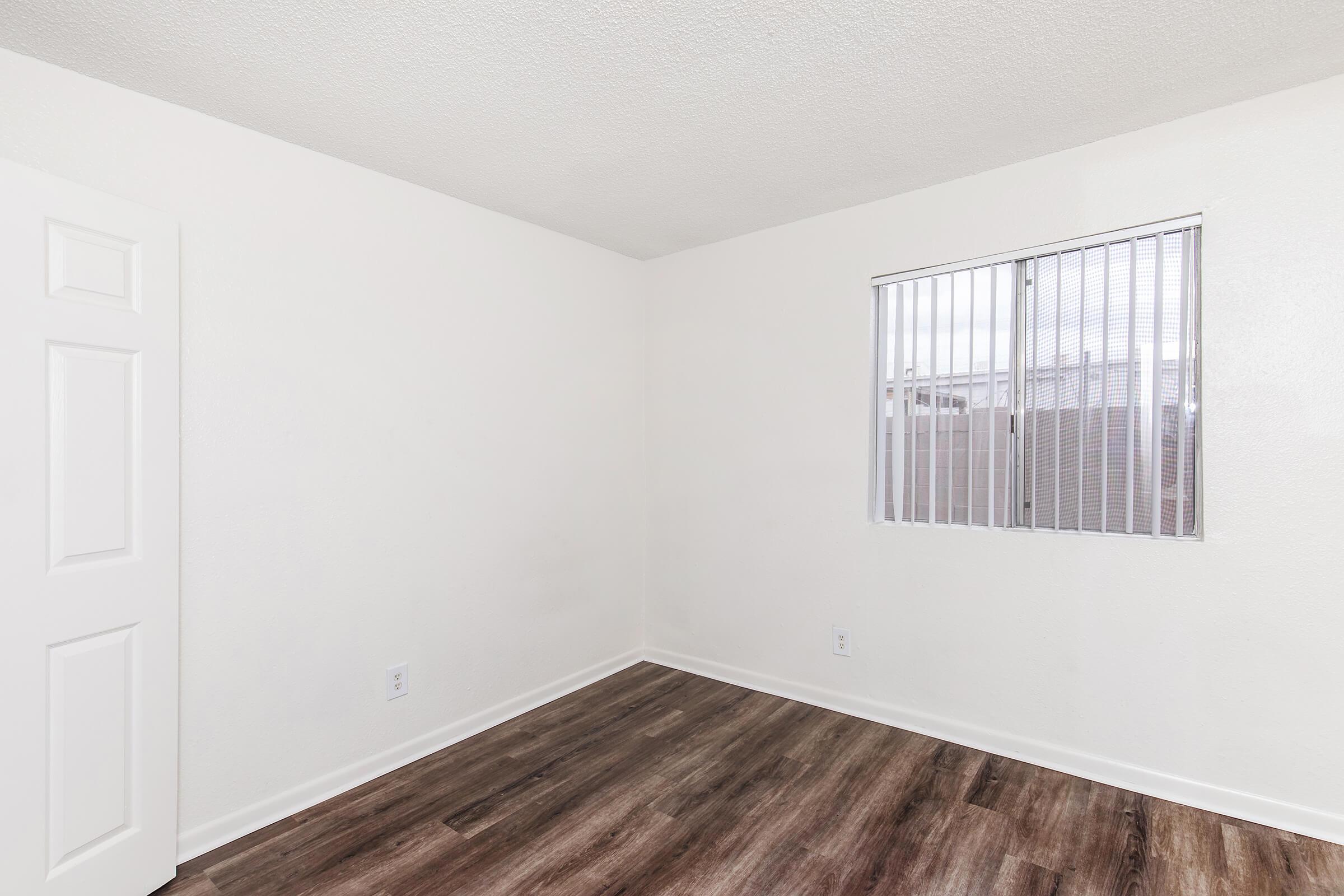 A vacant room with light-colored walls and a window featuring vertical blinds. The flooring is a dark wood-like laminate. A white door is visible on the left side of the image, and the overall atmosphere is bright and uncluttered.