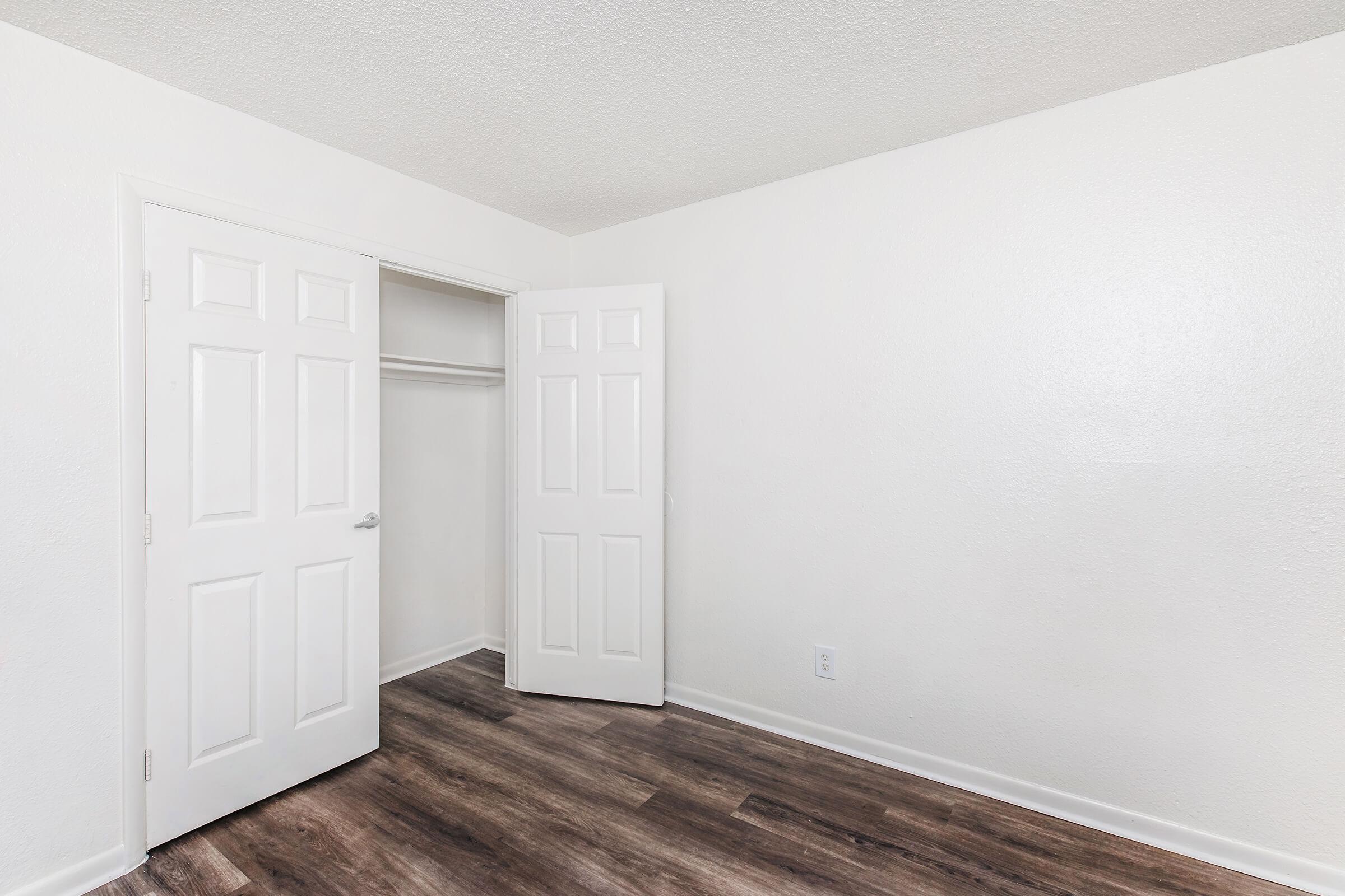 Empty room with light-colored walls and a wooden floor. Two white doors are open, revealing a closet space. The room appears well-lit and minimalistic, providing a spacious feel with no furniture or decorations.