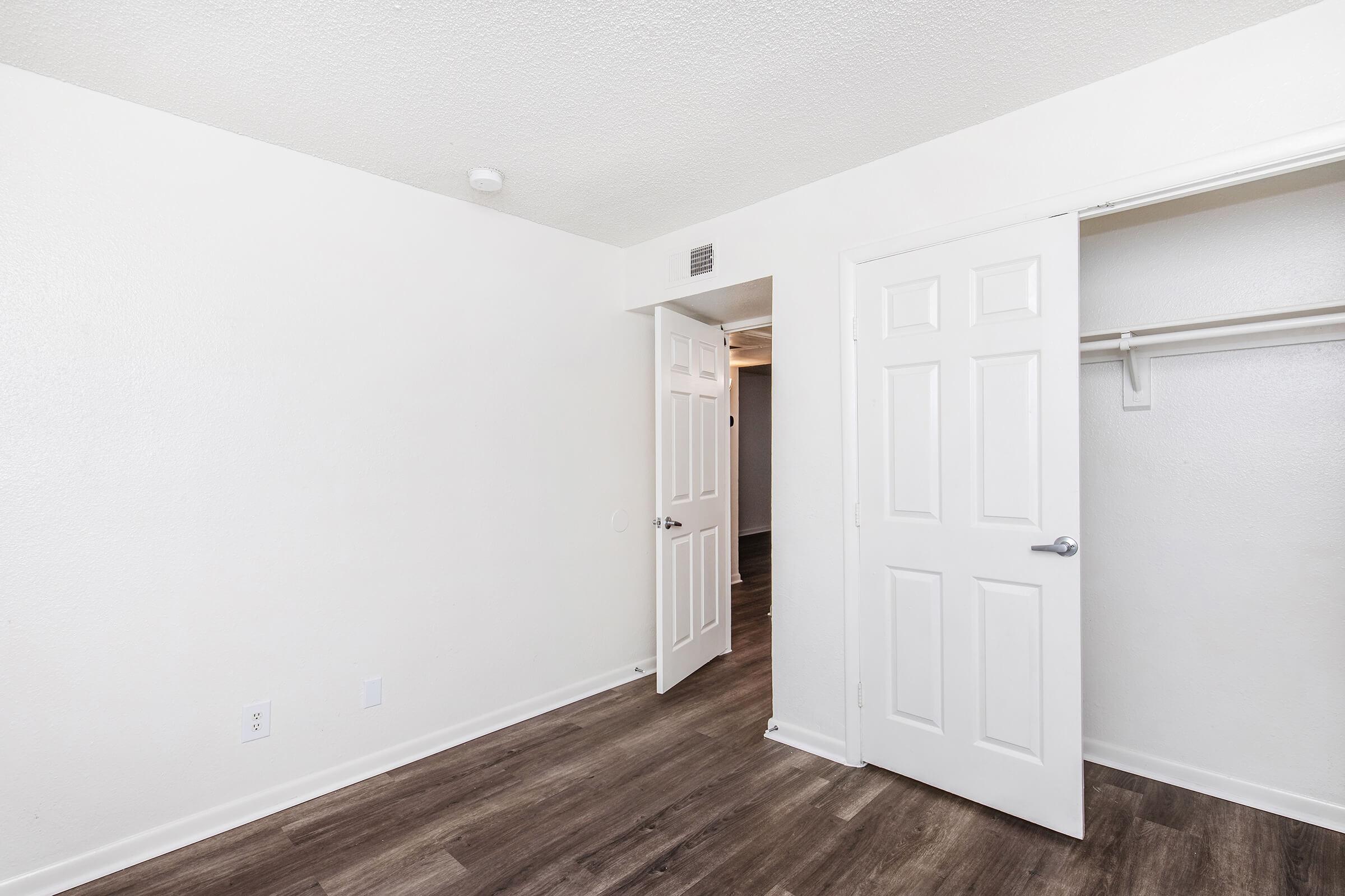 Empty room with light-colored walls, a wooden floor, and an open door leading to another space. There is a closet on one side with a hanging rod but no shelves. The lighting is bright, giving the room a spacious feel.