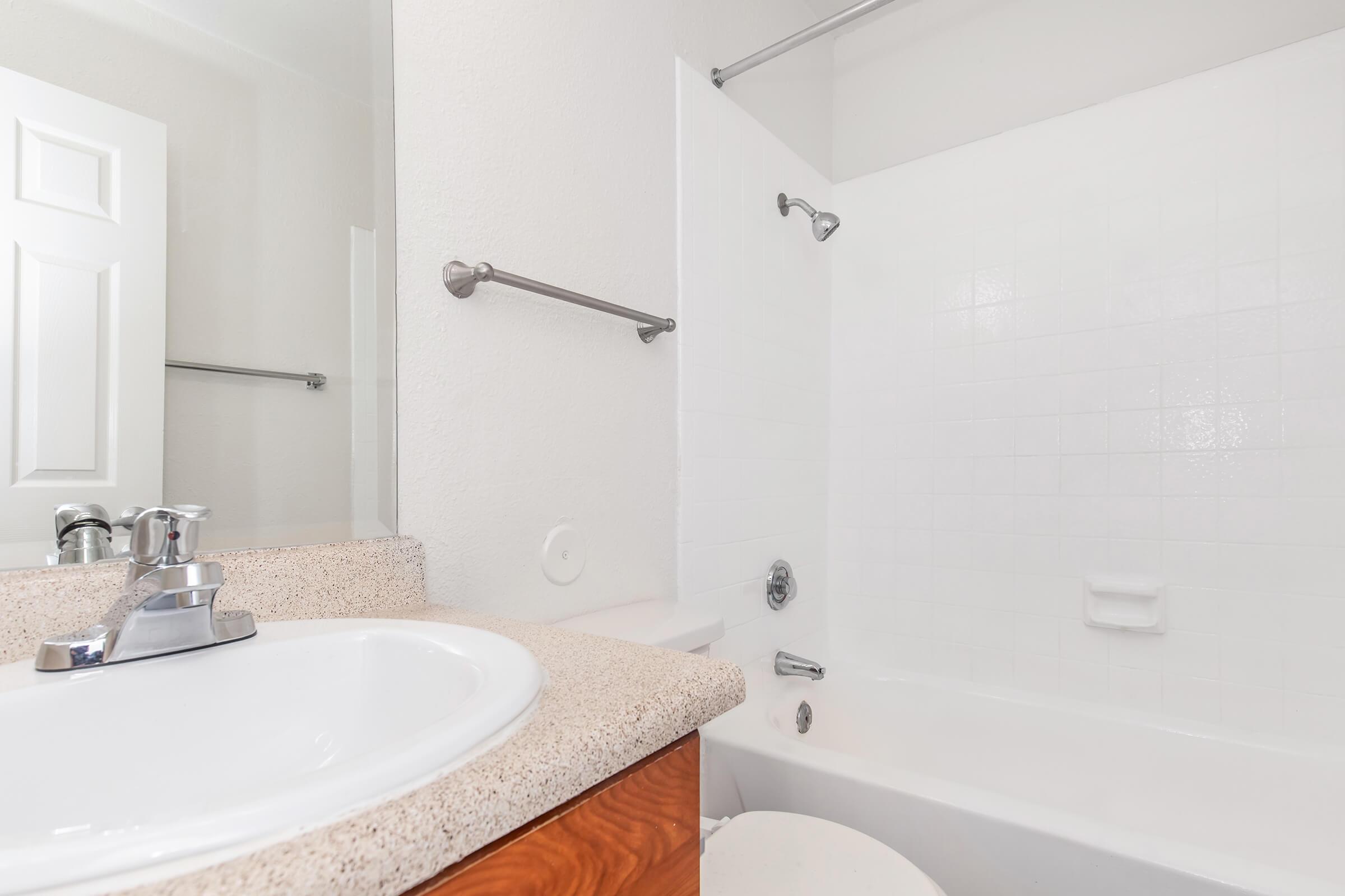A clean, bright bathroom featuring a white bathtub and shower combination, a sink with a faucet, and a mirror. The walls are painted white, and there's a towel bar mounted on the wall. The floor is not visible, and the overall design is simple and modern.