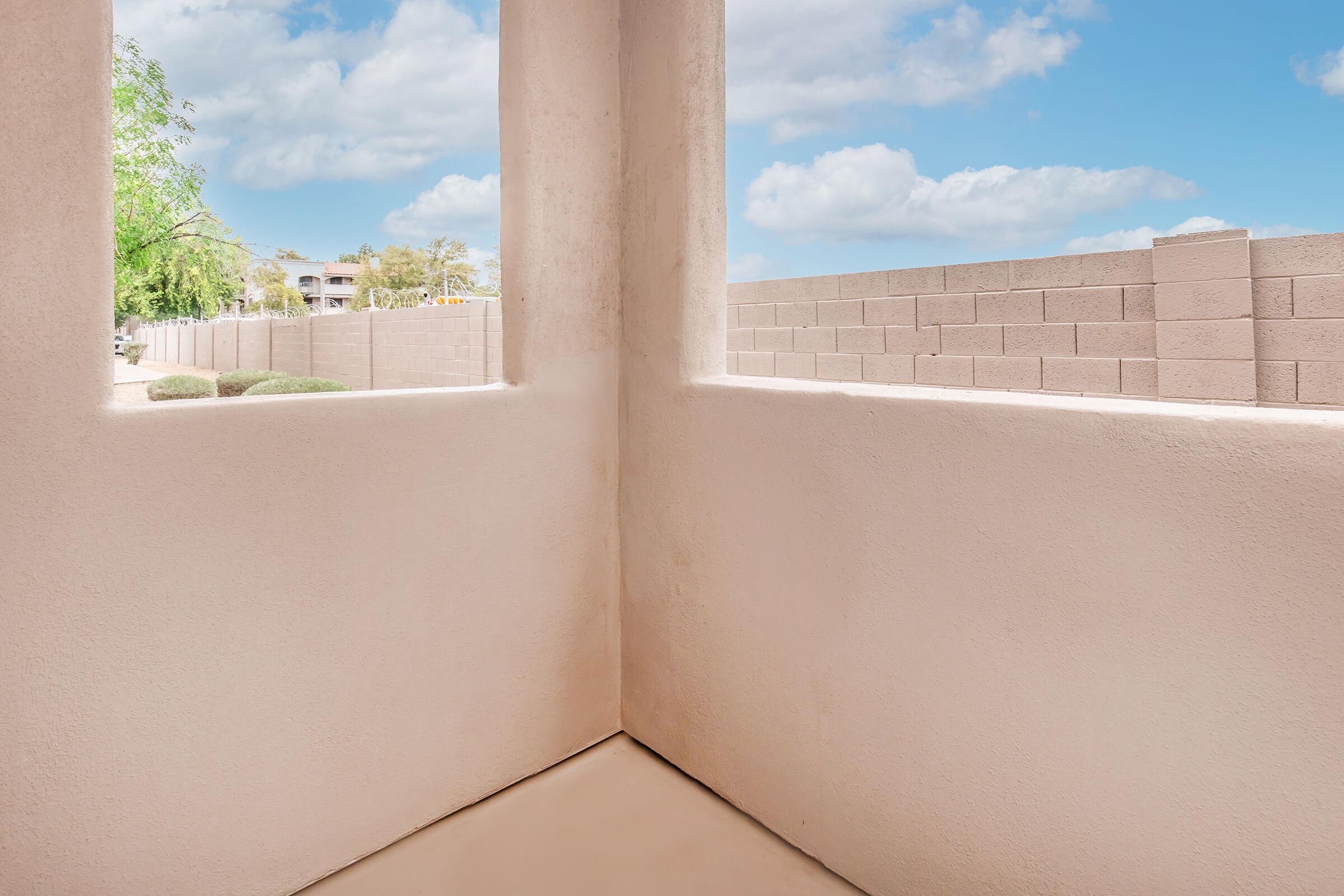 A corner view of a minimalist indoor space with light-colored walls and a clean floor. The scene features two open windows revealing a sunny outdoor landscape with some greenery and a block wall in the distance under a blue sky with fluffy clouds.