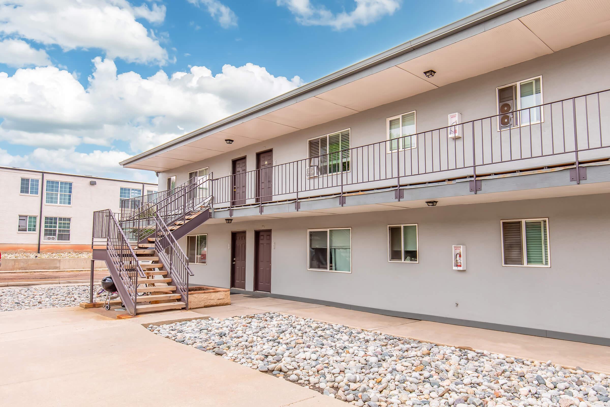 A multi-story apartment building featuring a stairway leading to the second floor, with several doors and windows. The exterior is gray, and the ground area has decorative gravel. The sky above is partly cloudy, enhancing the overall aesthetic of the property.