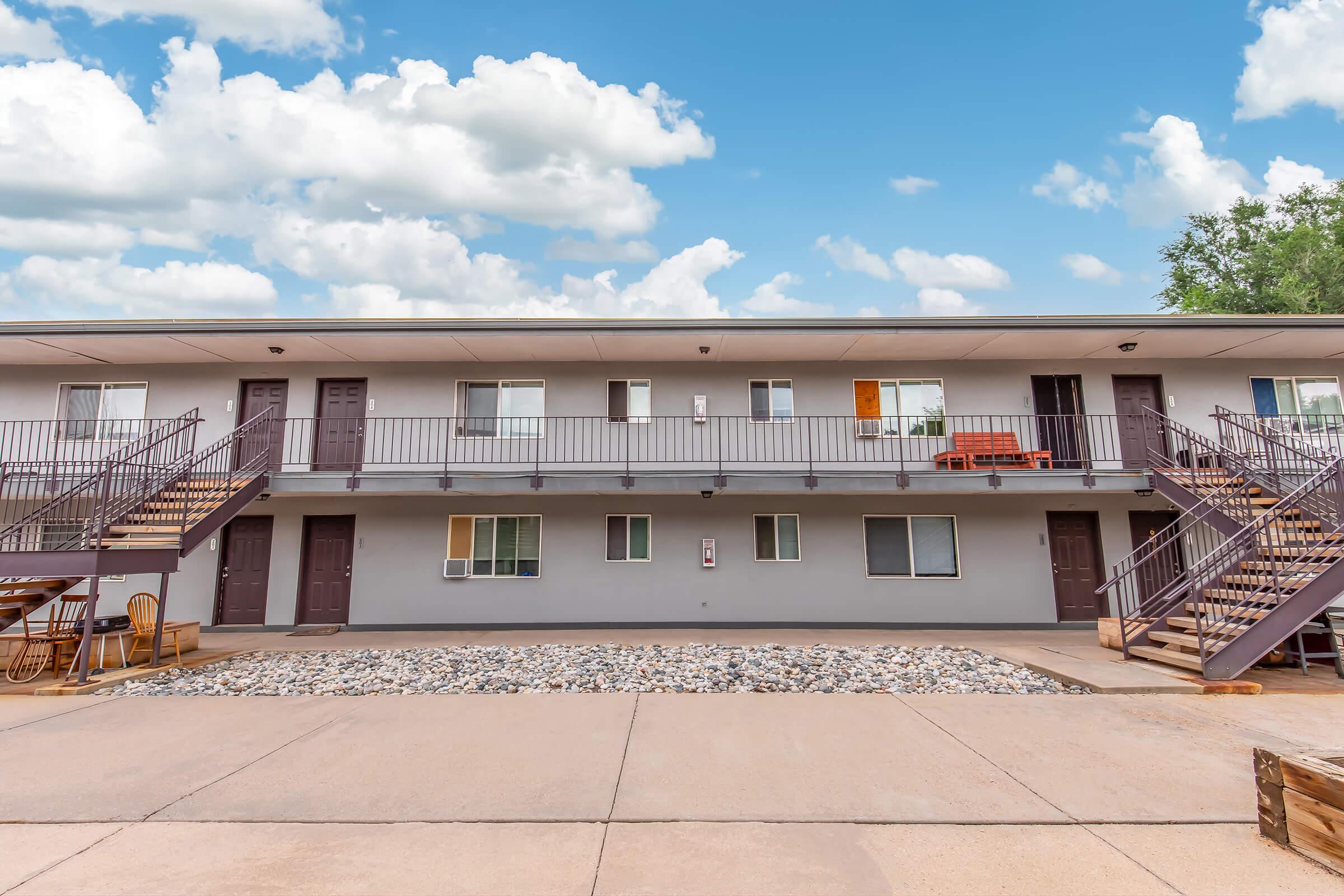Two-story motel-style building with multiple rooms, each having a door and window. Staircases lead to the upper level. There is a small courtyard area with stones and outdoor seating. The sky is blue with scattered clouds.