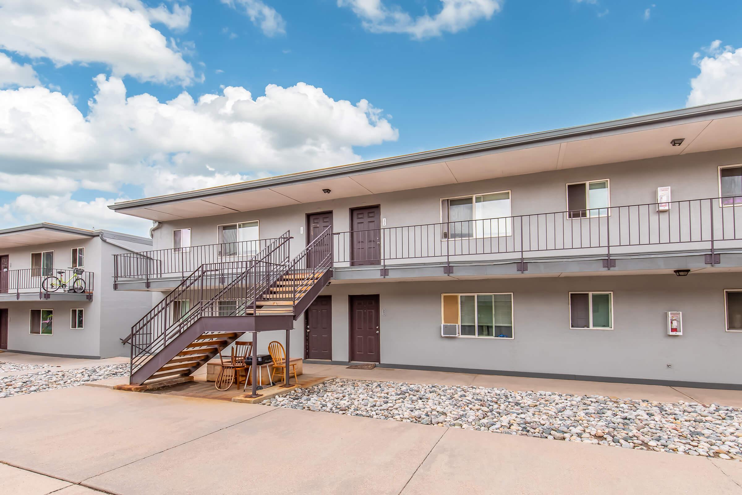 A two-story apartment building with a gray exterior, featuring a central staircase leading to the second floor. The ground level has multiple doors, and there are chairs set outside. A bicycle is mounted on the railing. The scene is set against a partly cloudy sky.