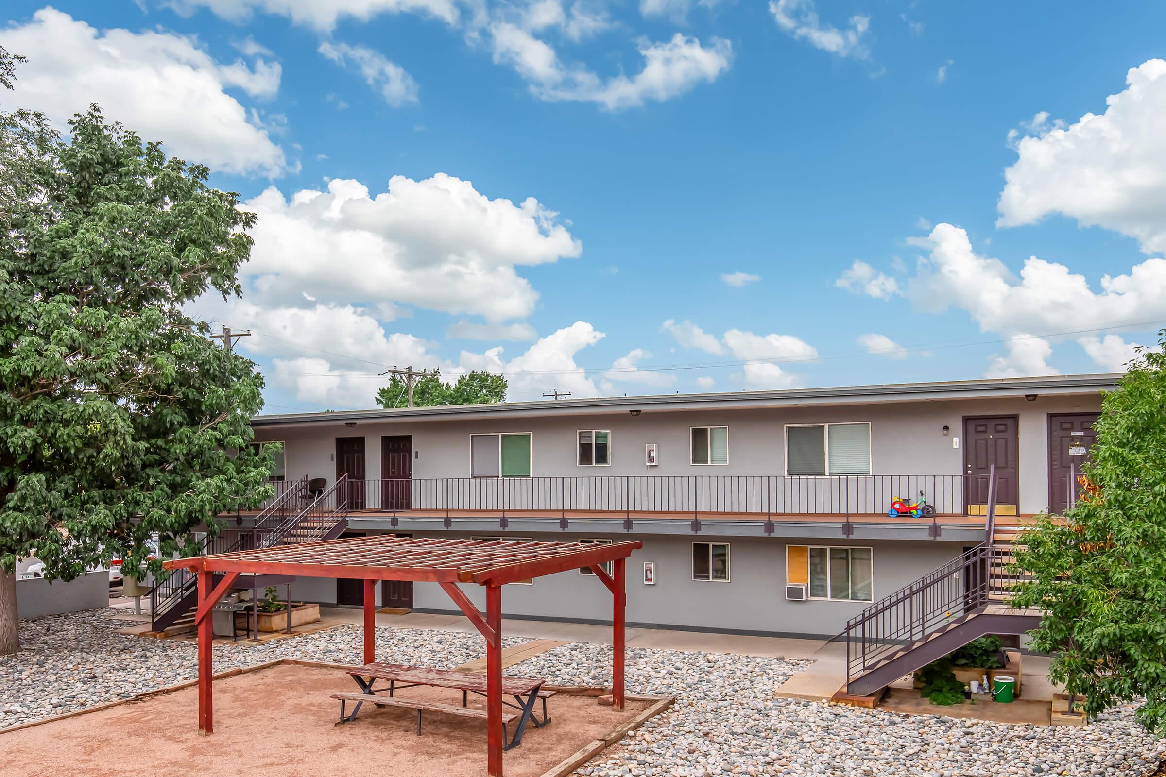 A view of a two-story apartment building with balconies and multiple units, surrounded by trees. In the foreground, there is a wooden pergola over a picnic table, with gravel and landscaped areas. The sky is partly cloudy, suggesting a sunny day.