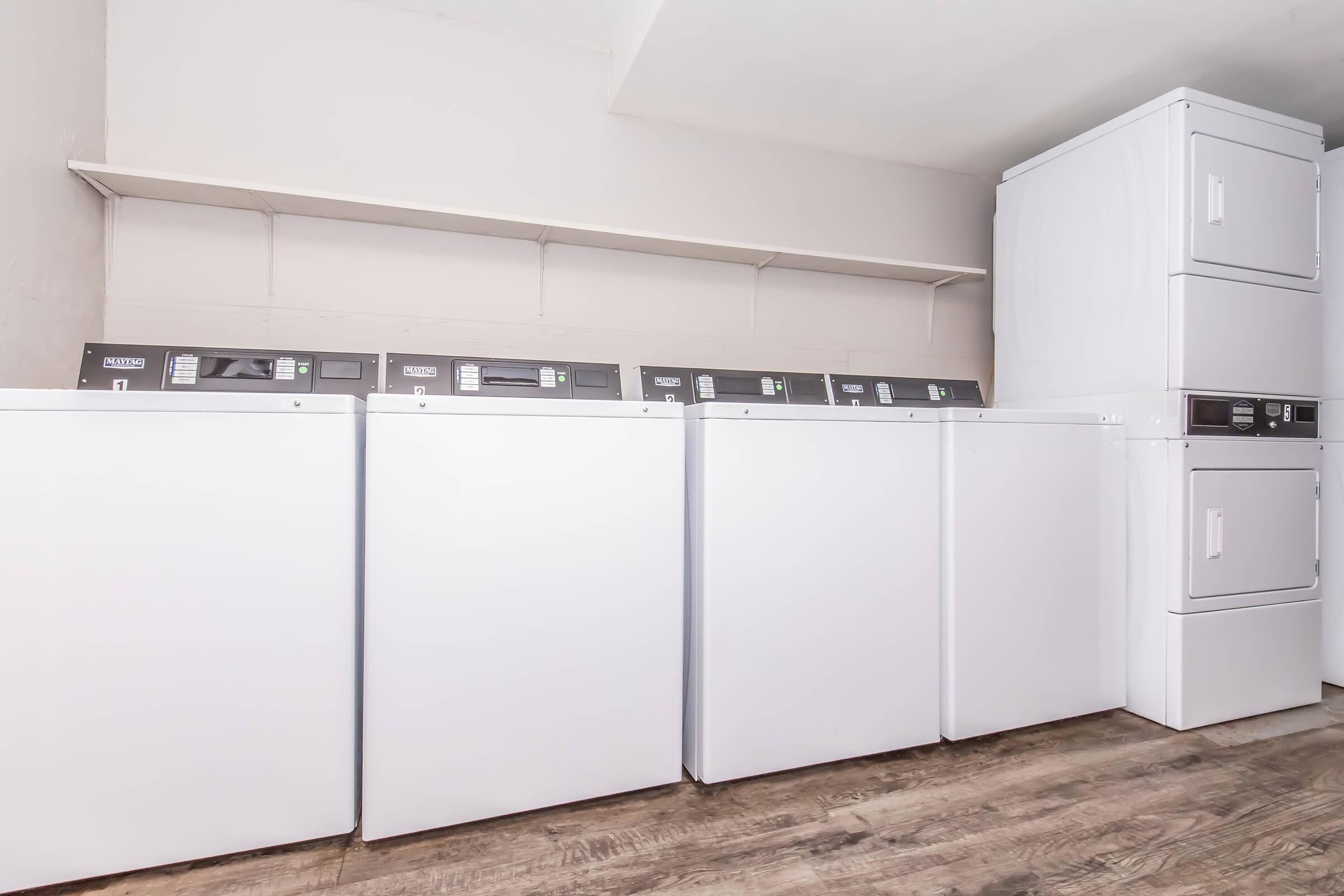 A laundry room featuring four white washing machines and a stacked dryer unit against a light-colored wall. There is a shelf above the machines, and the floor is made of wood. The overall space appears clean and organized, suitable for laundry tasks.