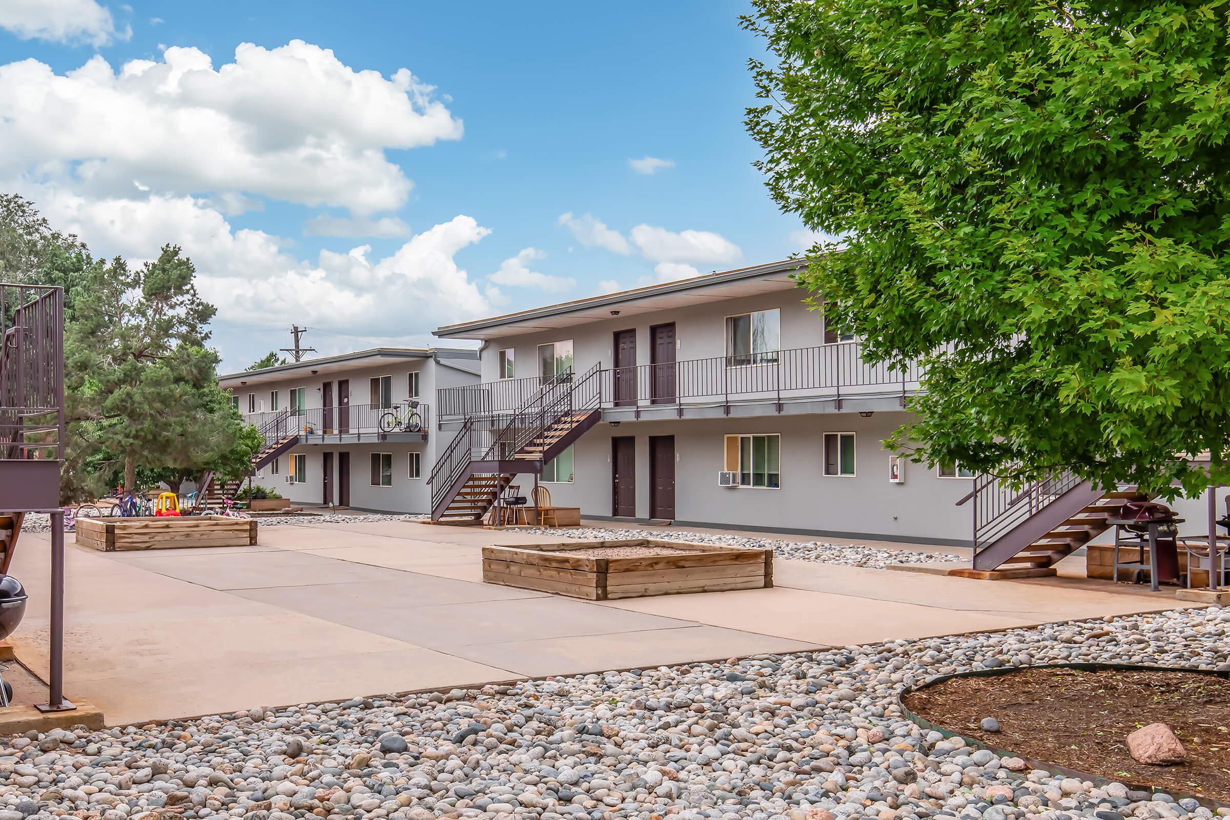 A view of a modern two-story apartment complex featuring multiple units with balconies. A central courtyard with a gravel pathway and wooden raised planters is visible. Lush green trees and fluffy clouds contribute to a pleasant outdoor atmosphere. Bicycles are parked nearby.