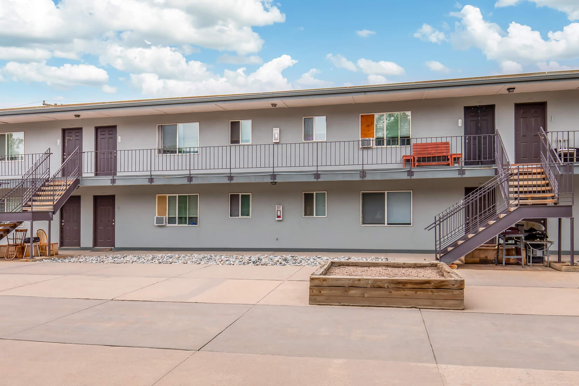 A view of a two-story apartment building with a gray exterior. The building features multiple doors and windows, a staircase on either end, and a small outdoor area with a raised wooden planter and a concrete surface. Light clouds are visible in the blue sky above.