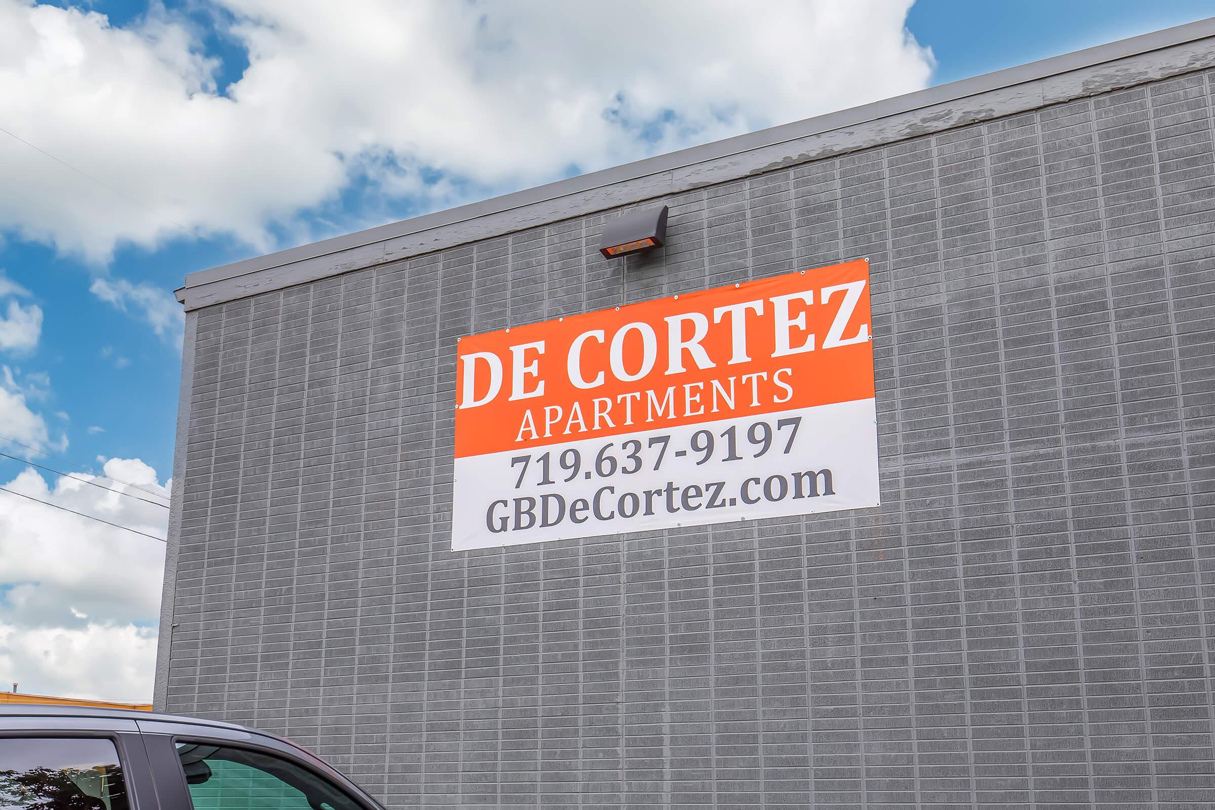 Sign for De Cortez Apartments featuring contact information: phone number 719-637-9197 and website GBDeCortez.com. The sign is mounted on a building with a cloudy blue sky in the background.
