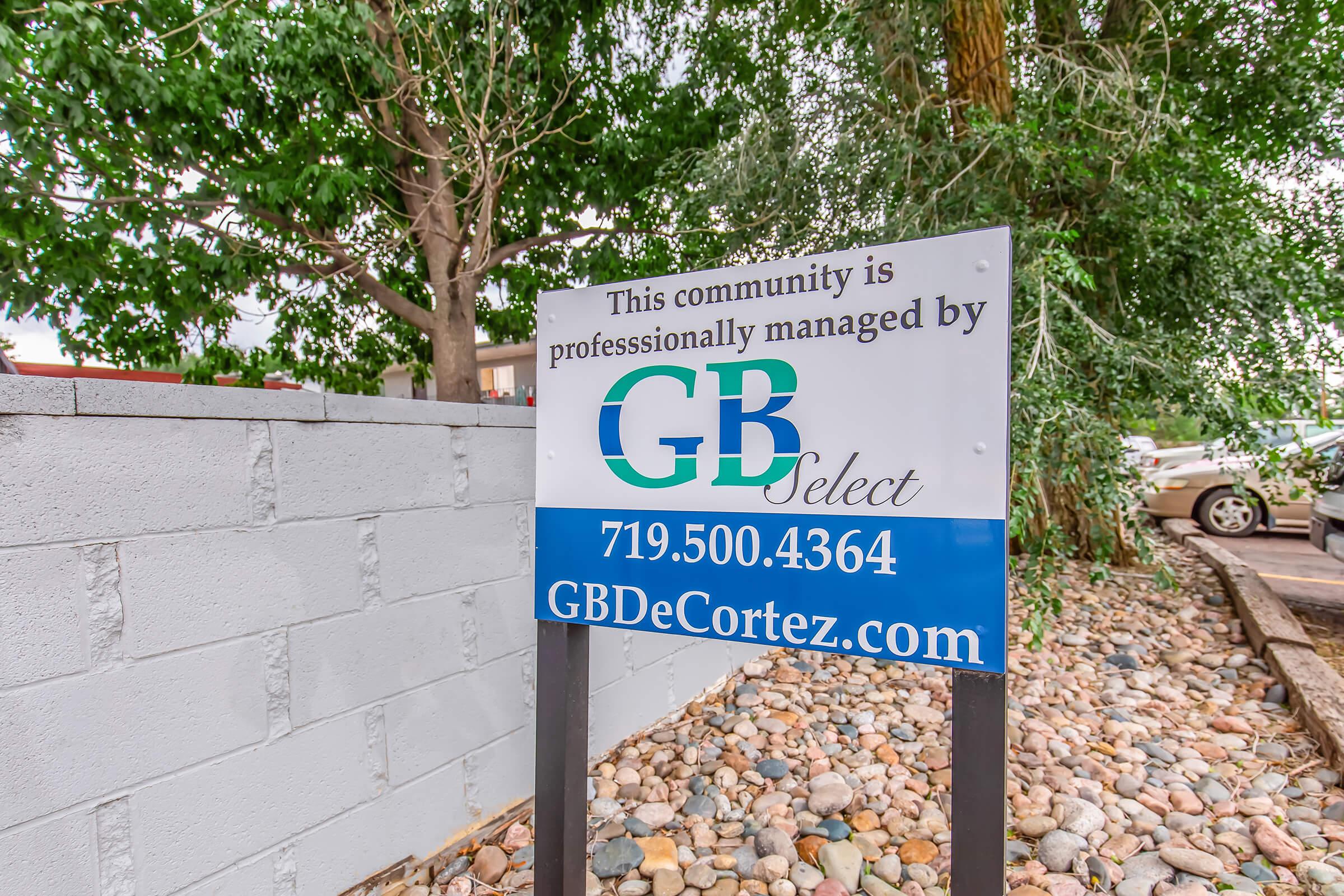 Sign indicating that the community is professionally managed by GB Select, featuring the company name, phone number, and website. The sign is placed near a landscaped area with trees and decorative rocks.