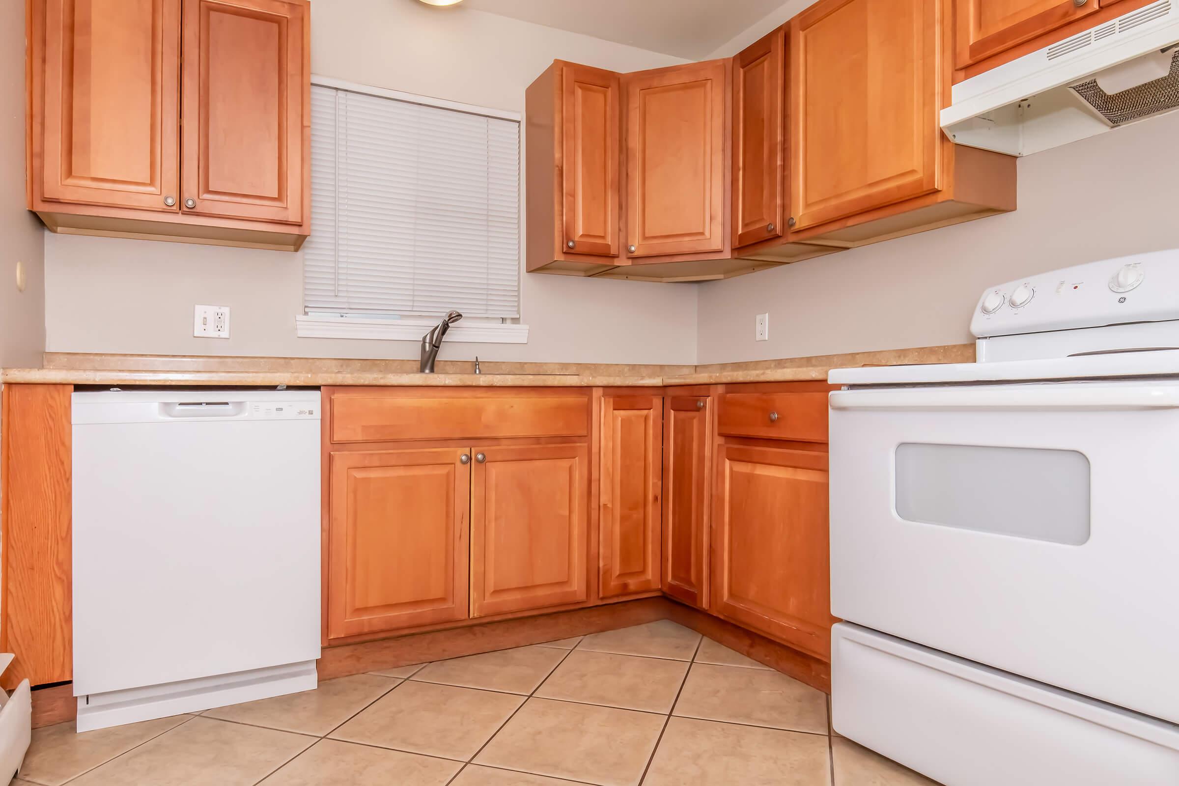 A modern kitchen featuring wooden cabinets, a white stove, and an integrated dishwasher. The countertop is beige with a tiled floor, and there's a window with blinds providing natural light. The space is organized and clean, emphasizing a functional cooking area.