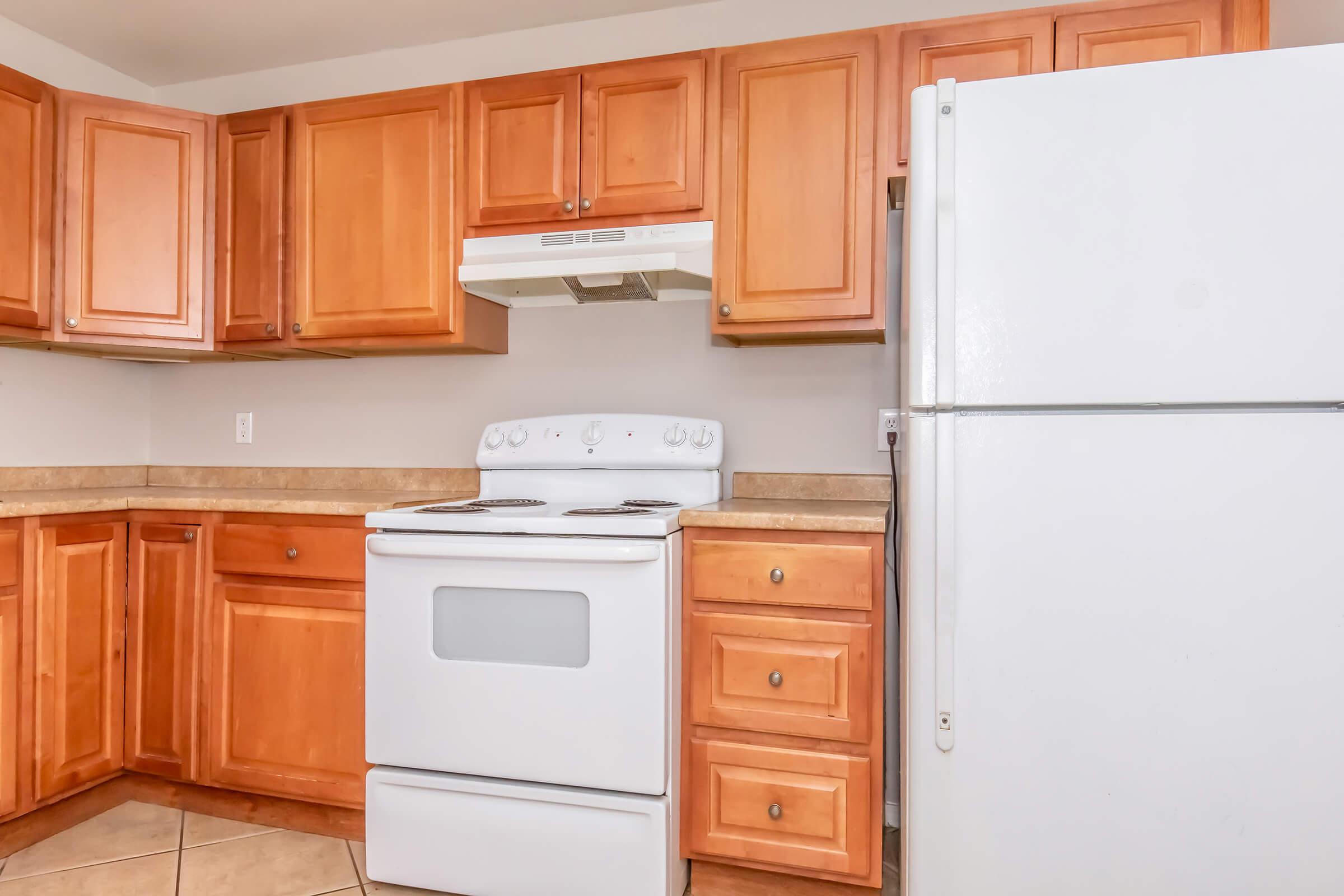 A kitchen featuring light wood cabinetry, a white stove and hood, and a white refrigerator. The countertop is beige and the floor is tiled. The space is bright and designed for cooking, with a functional layout.