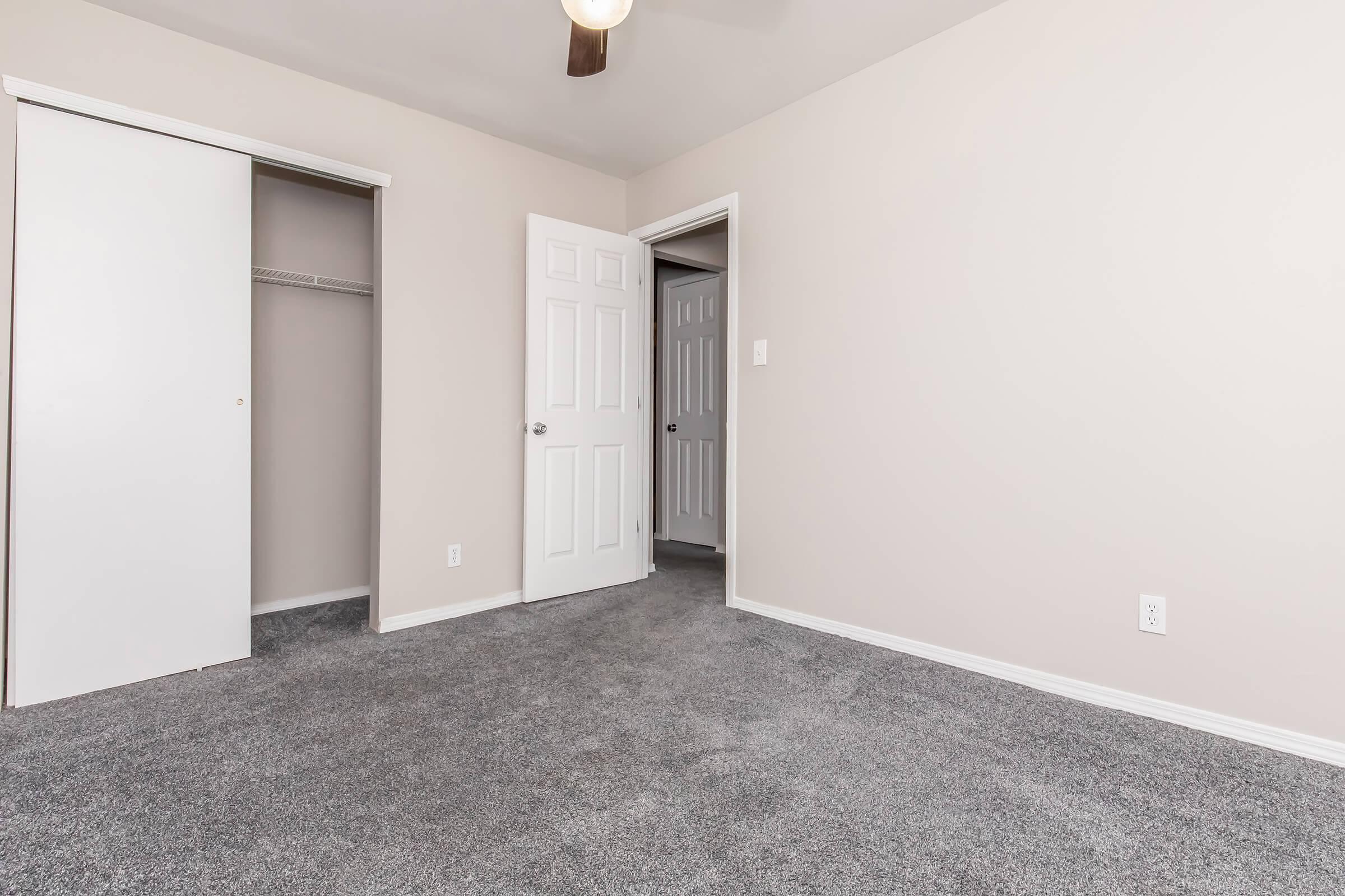 A spacious, empty room with light-colored walls and gray carpet. There are two doors: one leads to a closet with sliding doors, and the other opens into a hallway. A ceiling fan is visible above, and the overall atmosphere is bright and inviting.