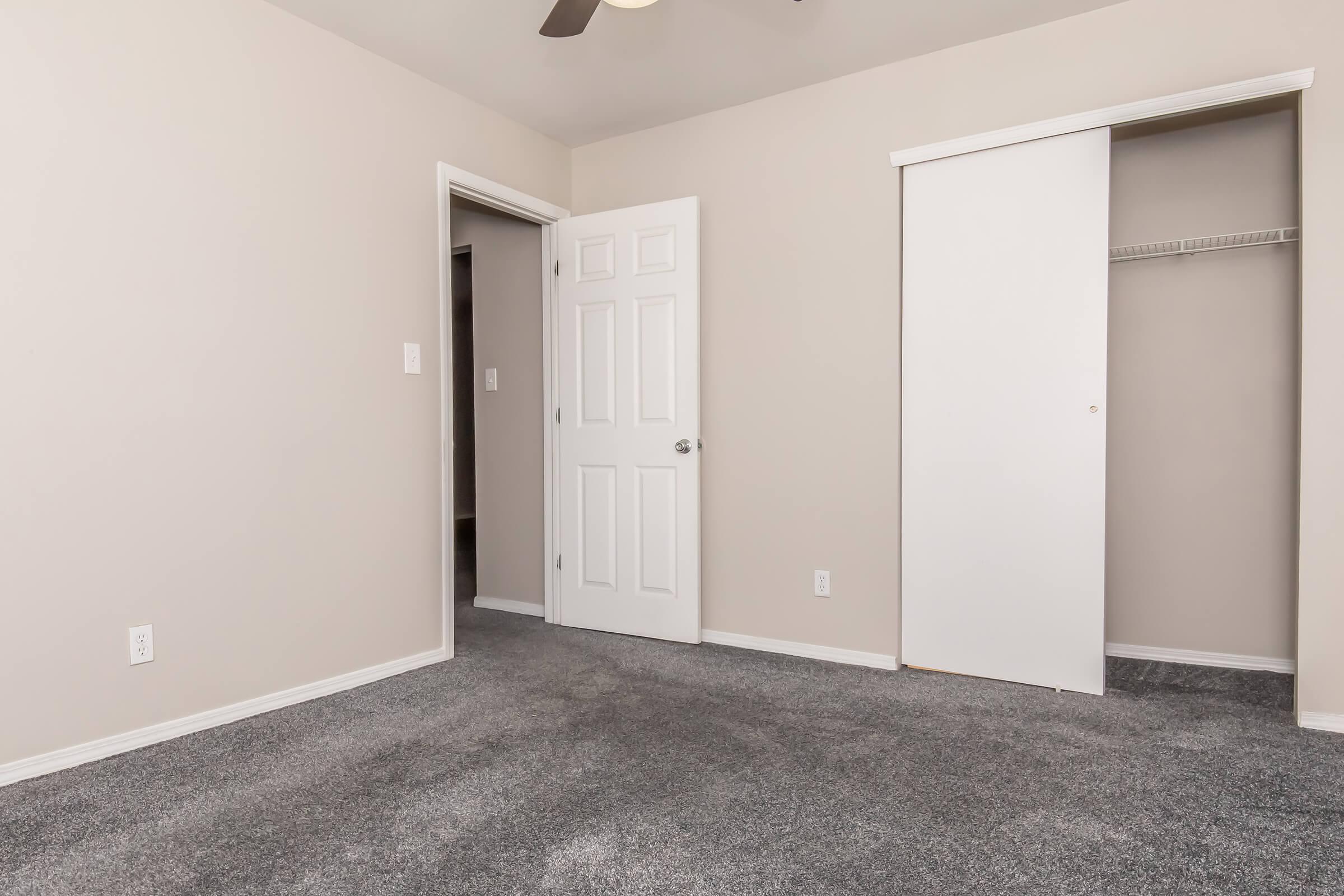 A simple, empty room with light gray carpeting and beige walls. It features a closed white door on the left, a closet with white doors on the right, and a ceiling fan. Natural light comes in from an unseen window, creating a neutral and spacious atmosphere.