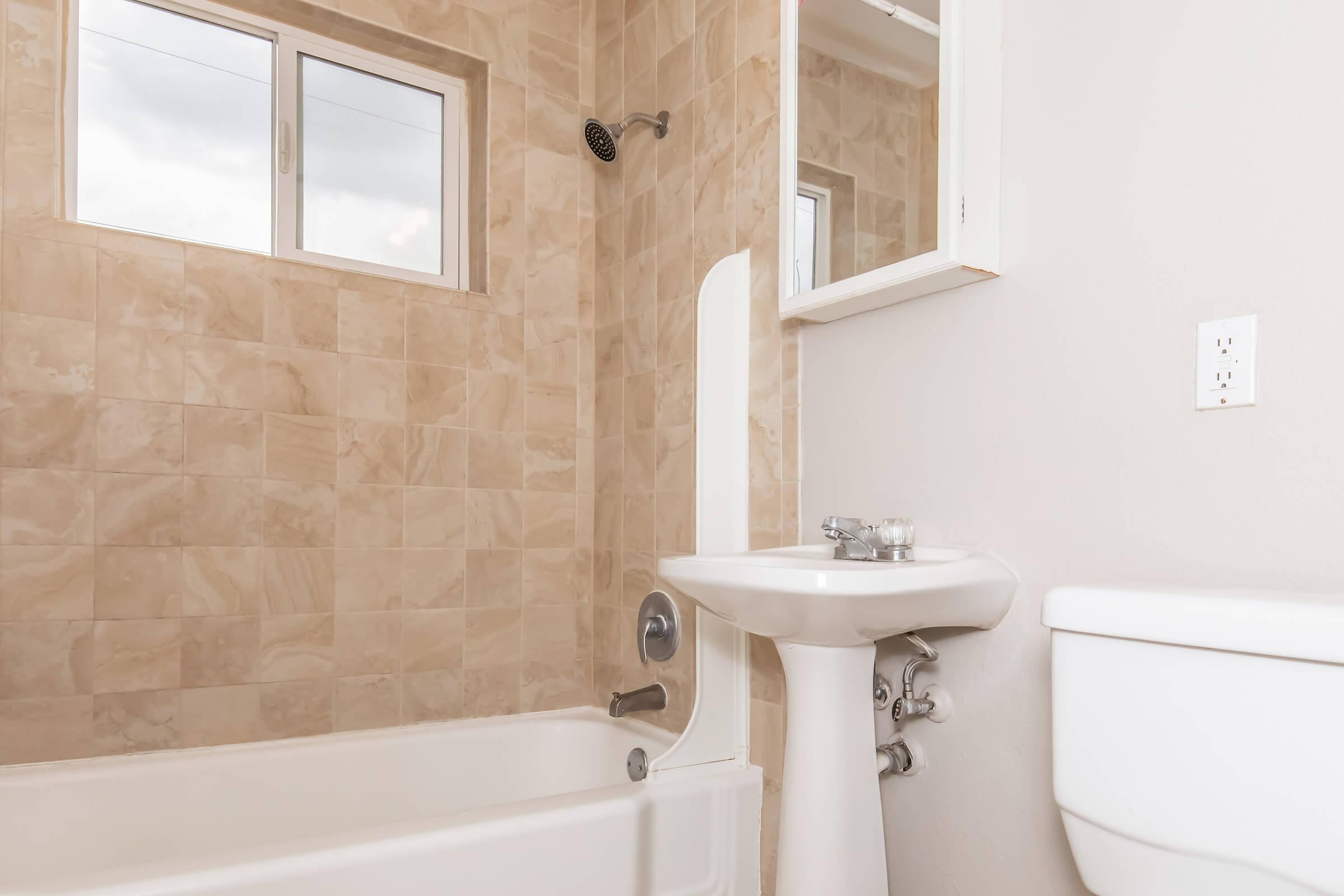 Bathroom interior featuring a beige tiled wall, a bathtub with a showerhead, a white sink with a mirror above it, and a white toilet. Natural light comes through a window, highlighting the clean and modern design of the space.
