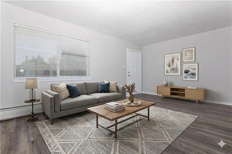 A modern living room featuring a gray sofa with decorative pillows, a wooden coffee table with a vase, and a side table with a lamp. There are two framed artworks on the wall, a light-colored area rug on the floor, and a window allowing natural light. The door to the outside is visible.