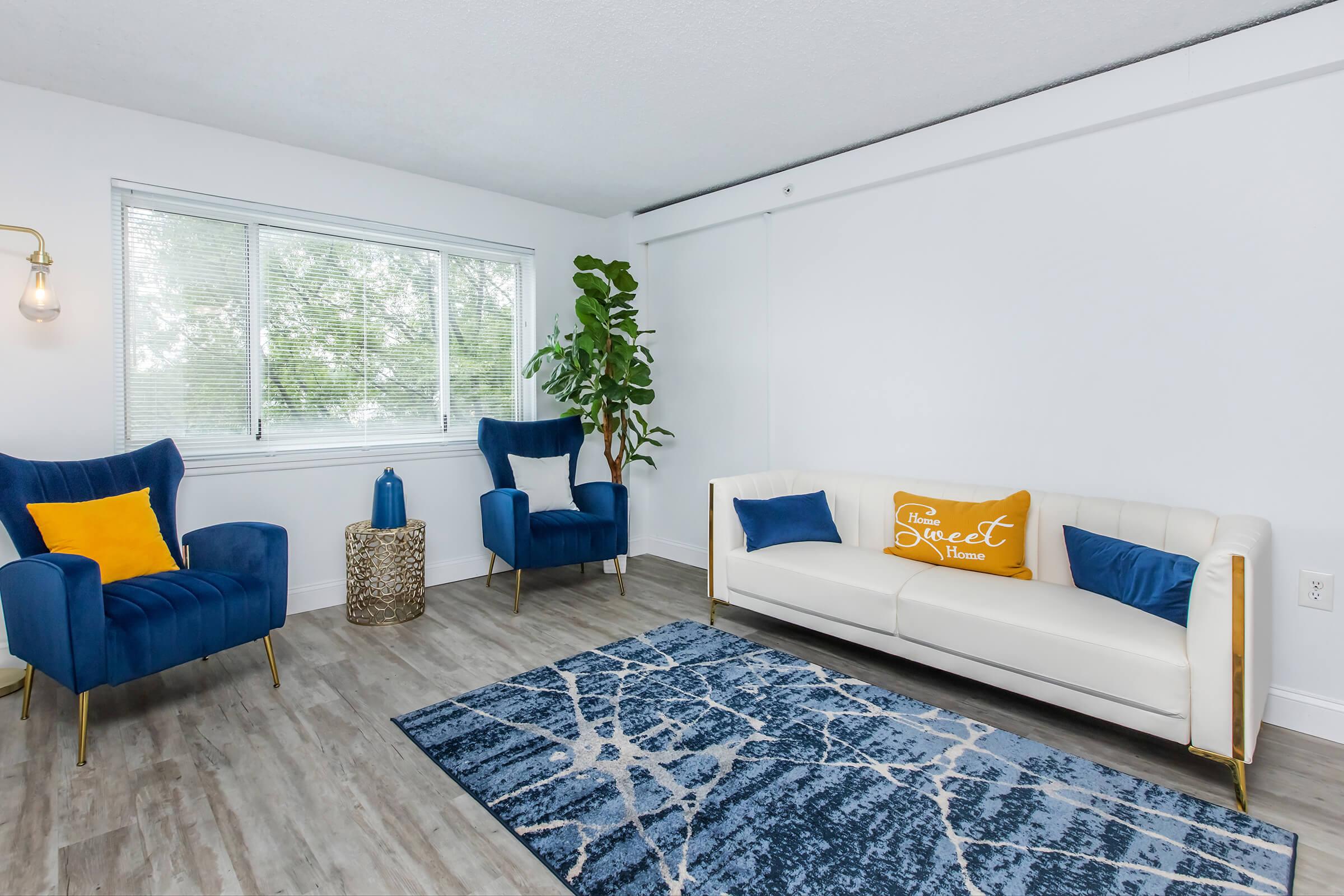 A modern living room featuring a white sofa with blue and yellow accent pillows, two blue velvet chairs, a textured area rug with a blue and white pattern, and a potted plant. Natural light comes through a window, and there’s a decorative basket on the floor, creating a cozy and inviting atmosphere.