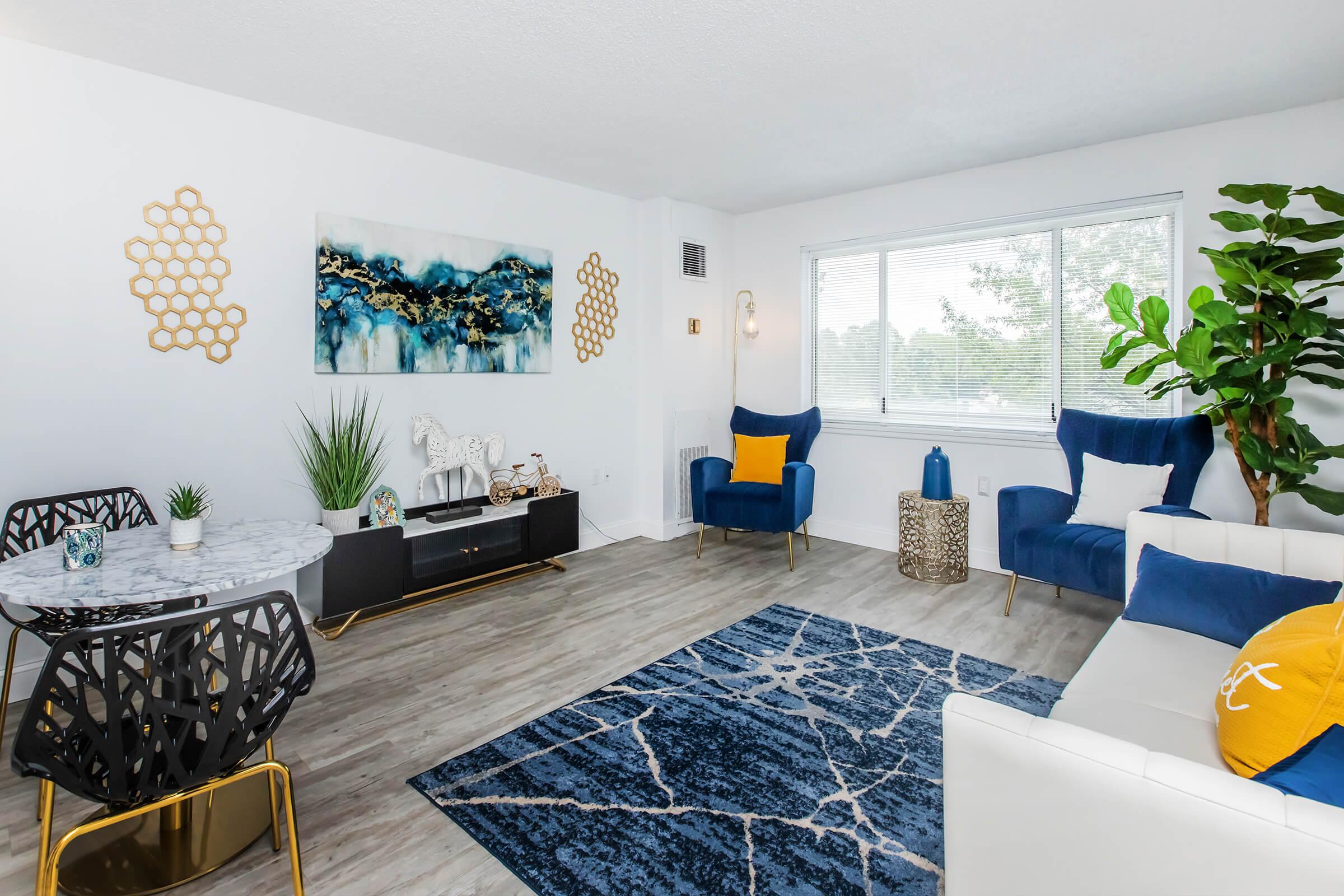 A modern living room featuring a blue and white color scheme. It includes a white sofa, two blue armchairs, a round marble dining table, and decorative gold accents. A large abstract painting hangs on the wall, and a potted plant adds greenery. A blue area rug with a geometric pattern covers the floor.