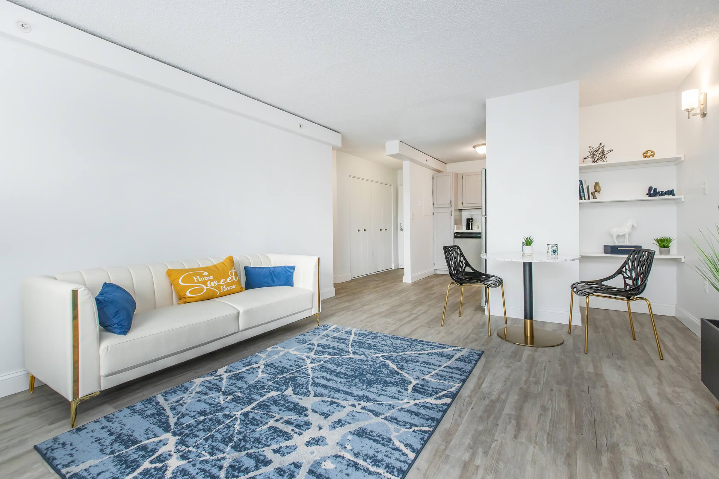 A modern, well-lit living space featuring a white sofa with blue and yellow pillows, a blue and white patterned rug, and a small dining table with two stylish chairs. The walls are painted white, and there are neutral-toned wood floors. A decorative shelf with minimalist items is visible in the background.
