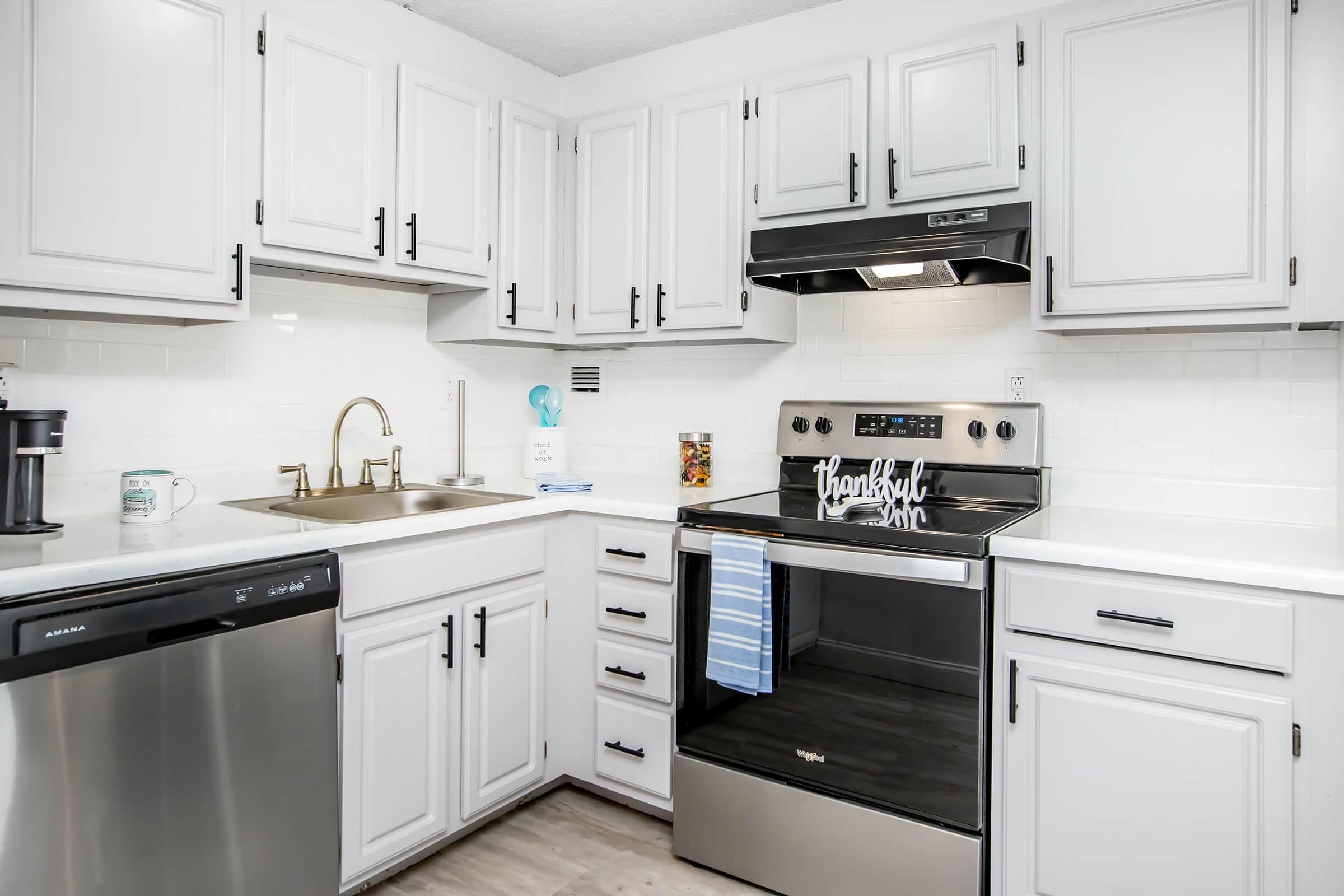 Modern kitchen featuring white cabinetry, a stainless steel dishwasher, and an oven with a glass stovetop. The sink area includes a gold faucet and there's a small decorative sign that says "thankful." The countertops are light and the space is well-lit, creating a clean, inviting atmosphere.